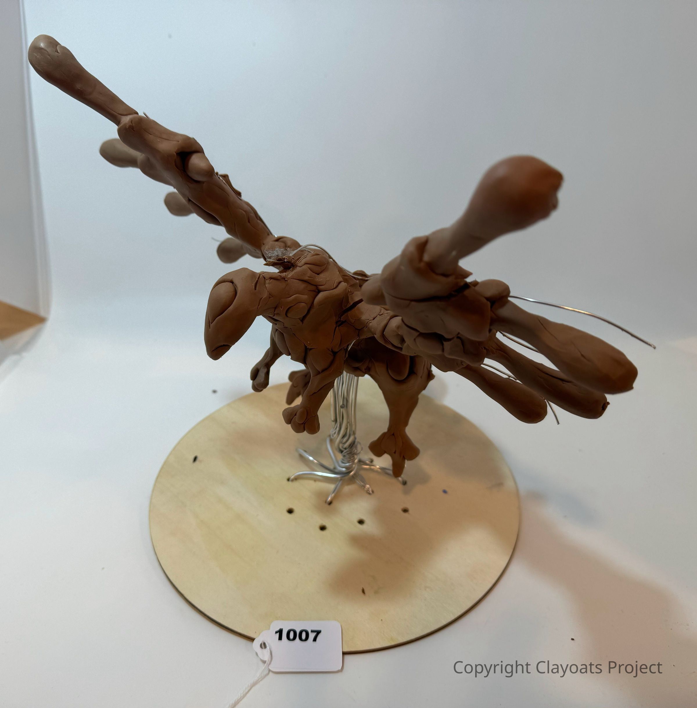 The image features a clay sculpture of a bird with its wings spread wide, perched on a circular wooden base. The sculpture's wings are textured and detailed, with some areas appearing rough and organic, giving the impression of movement. The base securely holds the figure with visible metal wires that help support the structure. The background is plain, highlighting the focus on the sculpture itself.