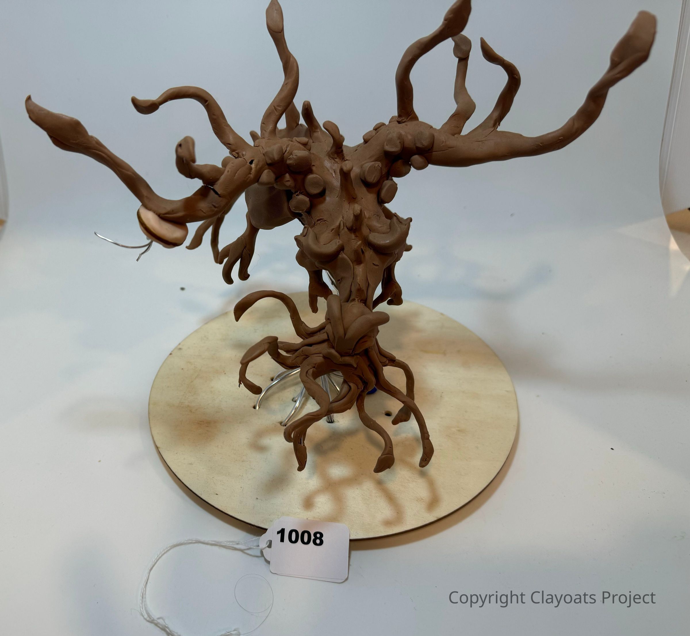 In the image, there is an intricately detailed clay sculpture of a fantastical tree-like creature with twisting, elongated branches extending upwards and outwards, while its roots seemingly curl and spiral beneath it. This imaginative sculpture, crafted in a brownish hue, sits on a circular wooden base, which supports its wiry structure. The texture of the clay exhibits a smooth yet uneven surface, highlighting the artist's ability to capture organic forms in an abstract approach.
