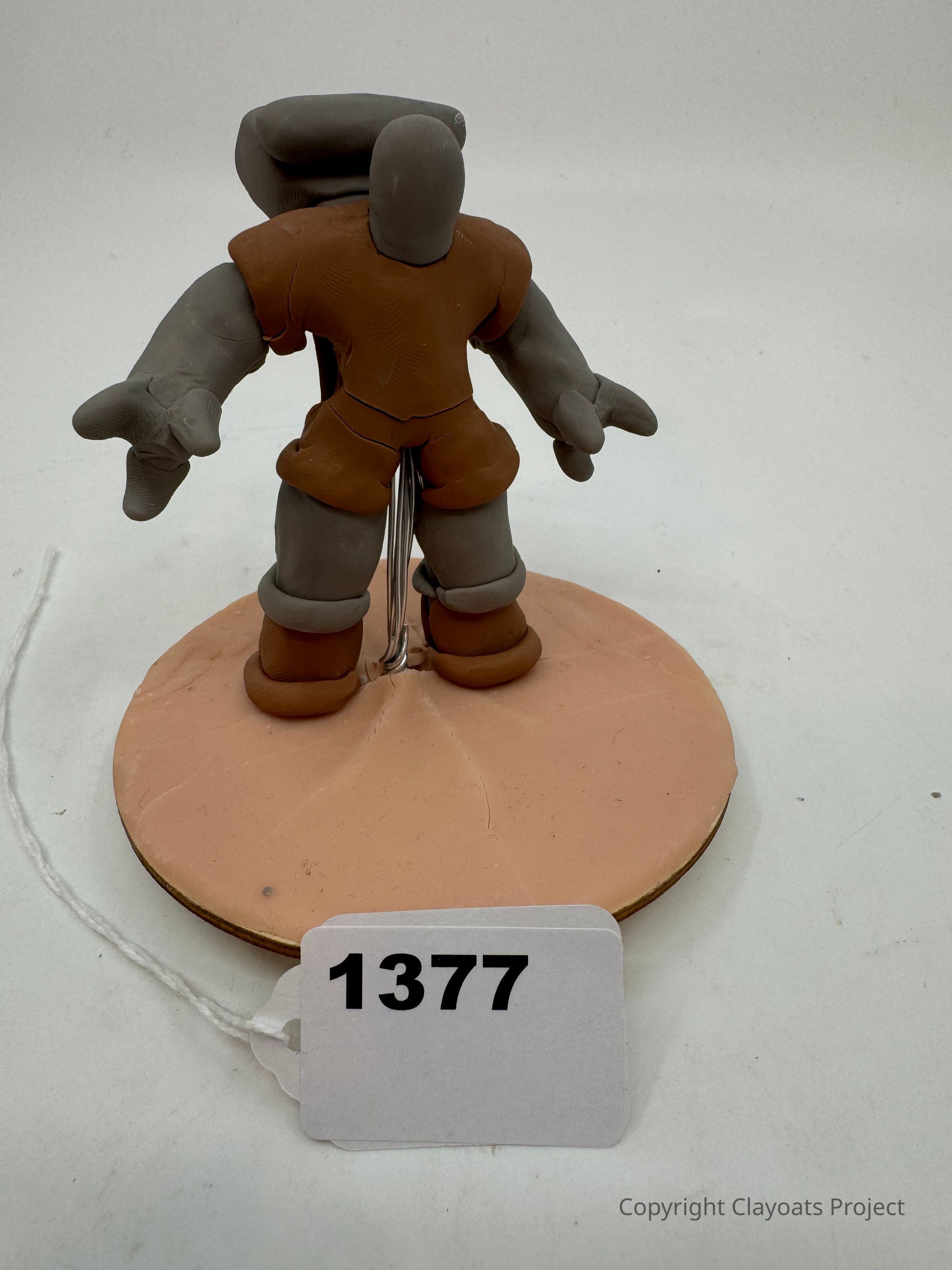 The image shows a small clay figurine resembling a simplified humanoid character, featuring exaggerated limbs and a smooth, featureless face, standing on a round base. Its body is composed of two colors: brown for the torso and feet, with gray for the head, arms, and legs. The figure has wire supports,