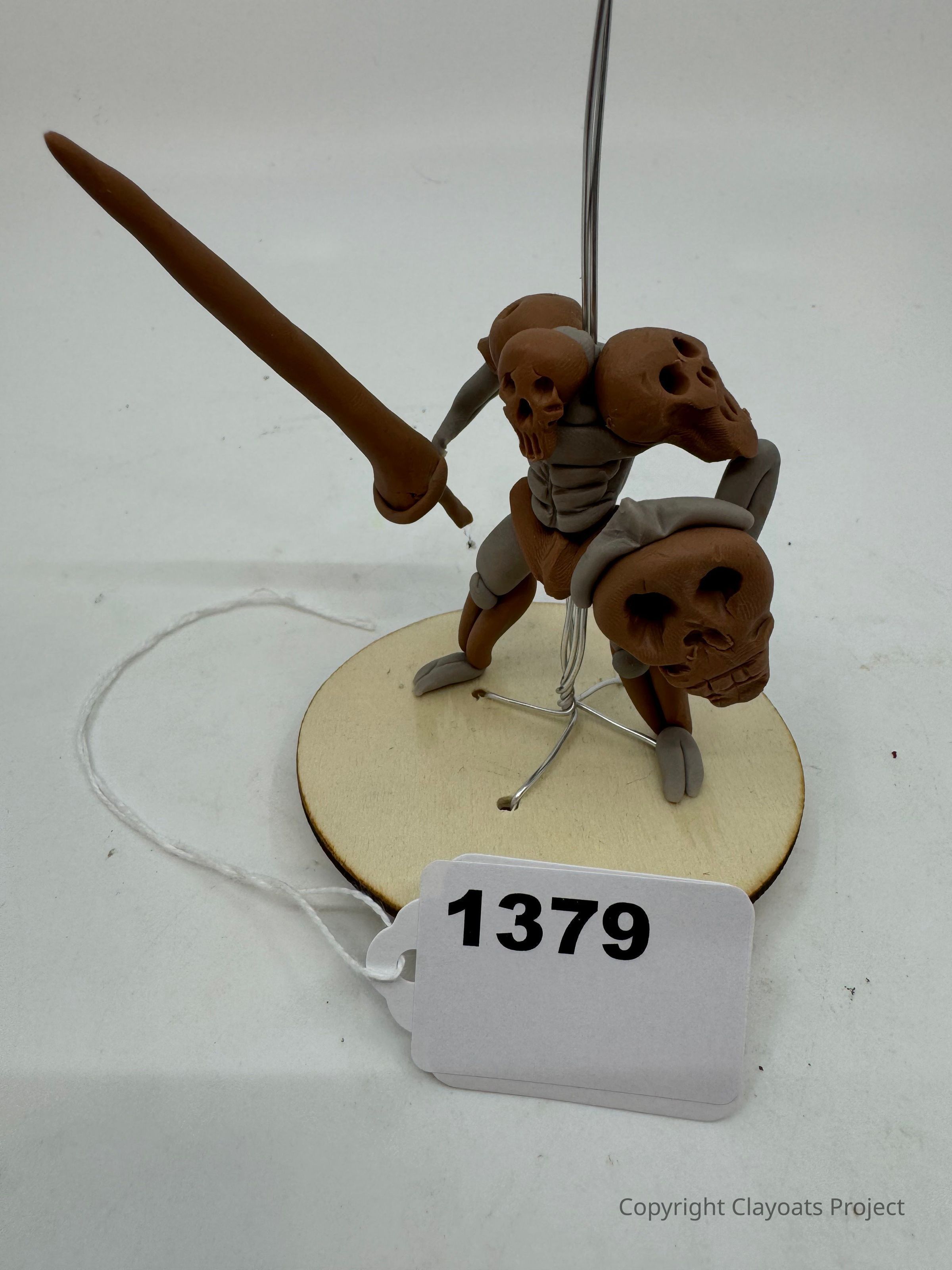 The image features a detailed clay figure resembling a skeleton warrior, with prominent skull motifs integrated into its armor, holding a long, brown sword in its right hand while standing on a round wooden base. The background is plain and neutral, accentuating the handcrafted model's intricate design elements and its earthy color palette of browns.