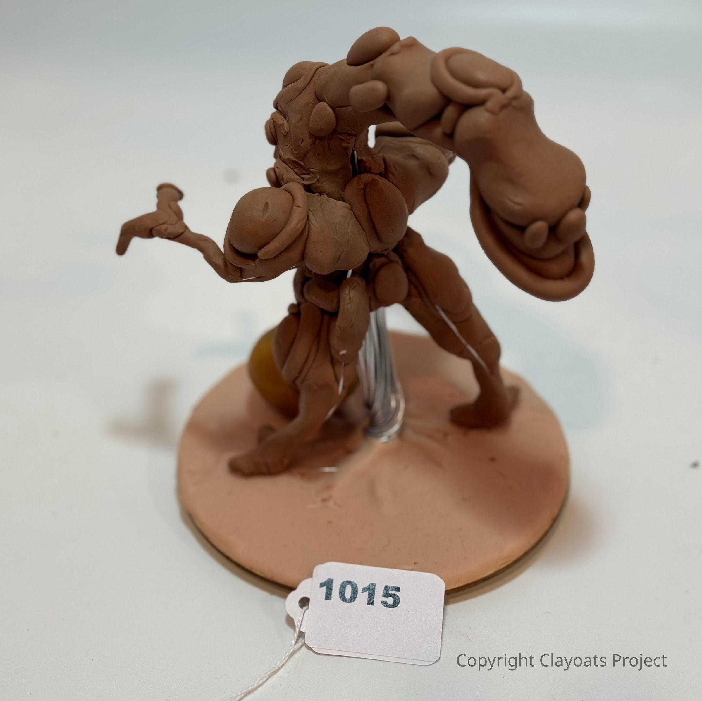 The image shows a small, abstract clay sculpture with an intricate, organic design mounted on a circular base, featuring a variety of rounded shapes and flowing lines, resembling a futuristic or fantastical creature. The sculpture is primarily a reddish-brown color, and it sits atop a flat, beige surface. The background is a soft white or light gray, giving the sculpture an exhibit-like appearance, and casting subtle shadows around the piece.