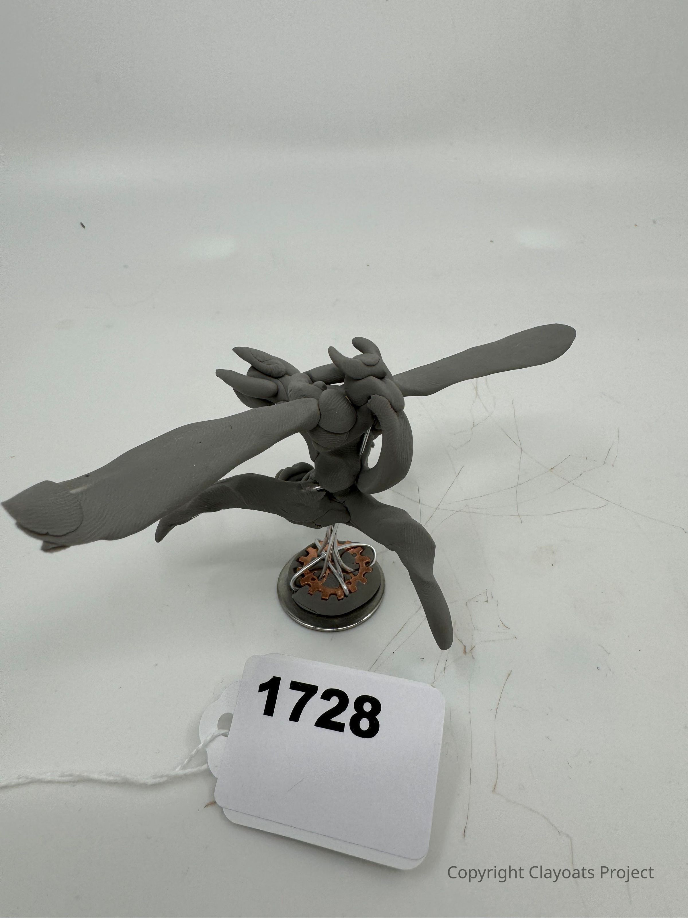 The image shows a sculpted model of a grey creature with elongated limbs, perched on a circular metal and gear base. Its surface appears textured, and it is positioned in a way that suggests motion or dance, with its wings or arms extended. The background is a plain, light-colored surface, creating a neutral setting that emphasizes the figure's distinct form and intricate details