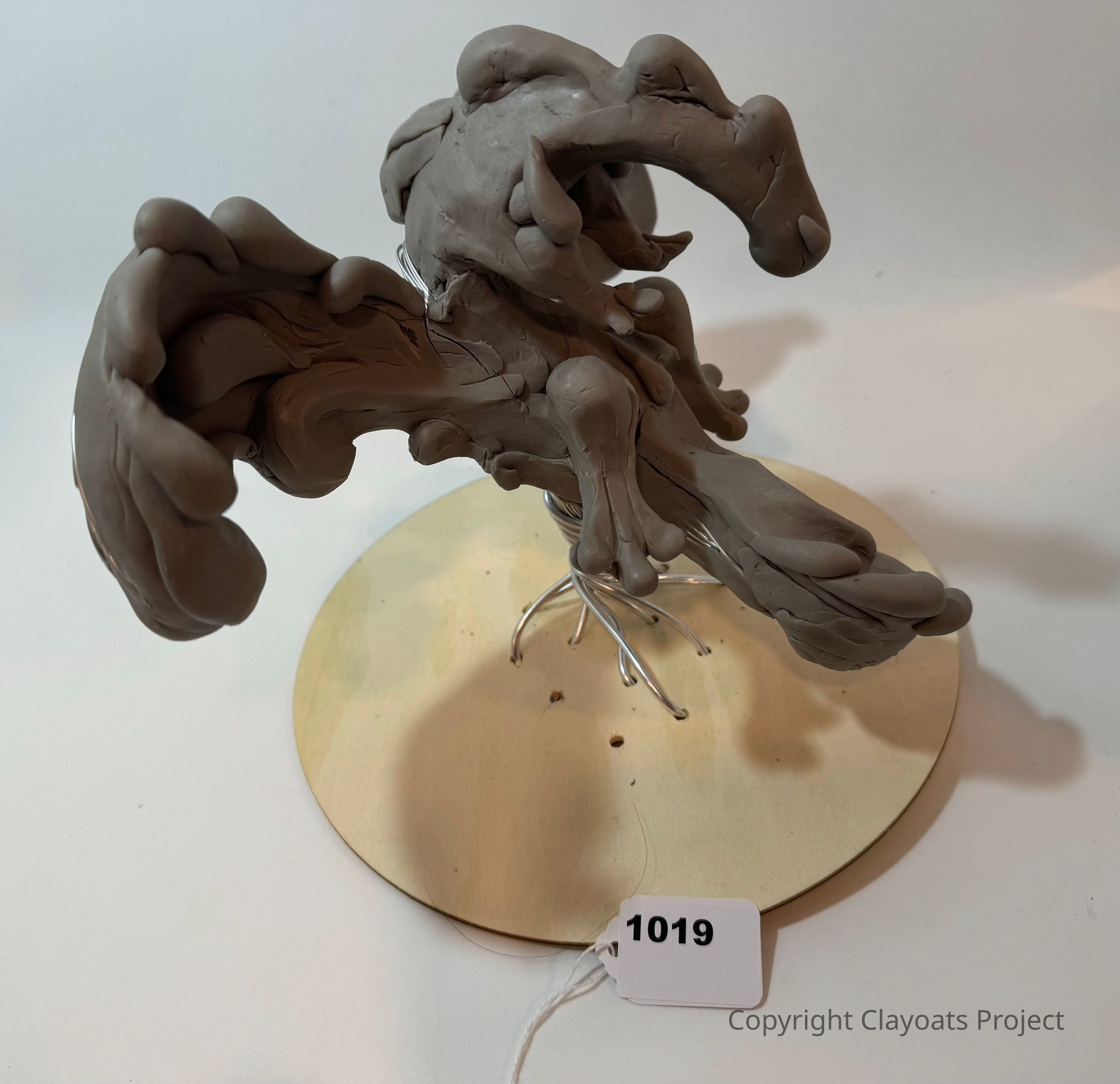 The image shows a sculptural artwork made from clay, consisting of abstract, swirling shapes that create a dynamic form mounted on a circular wooden base; the clay features organic, tendril-like extensions that seem to radiate outward, giving the piece a sense of fluidity and movement, while the base supports the structure with visible wire armature providing stability and balance, each contour appearing to undulate and curl, conveying an impression of continuous transformation; the sculpture rests prominently on the wooden base.