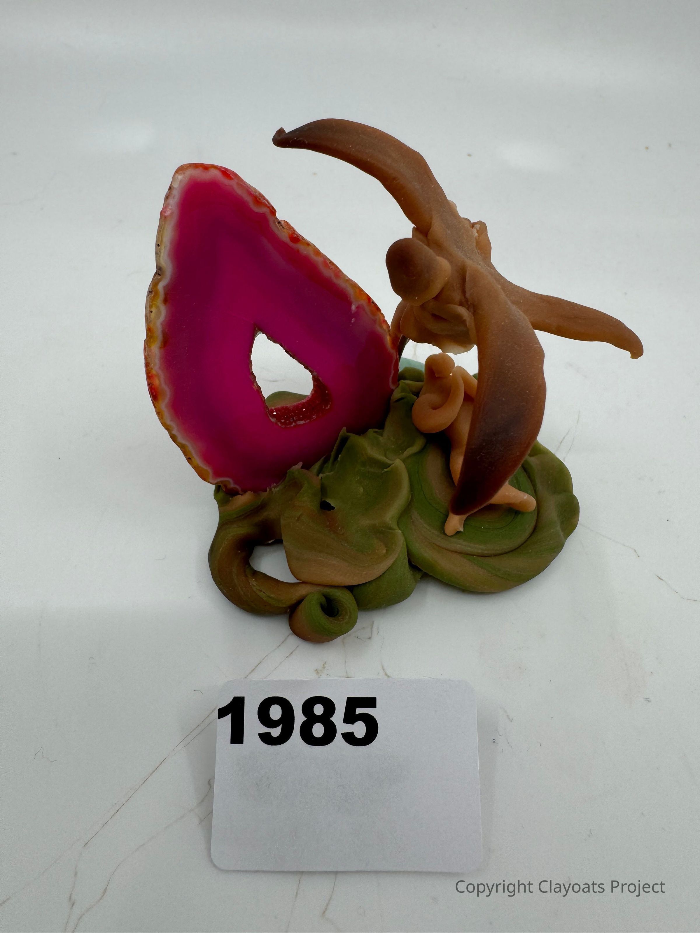 The image features a creative clay sculpture of a figure with outstretched arms, facing a vibrant pink and agate-like geode slice. The figure is positioned on a swirl of earthy green and brown clay, suggesting a natural environment. The backdrop is plain, ensuring the focus remains on the intricate details and vibrant colors of the sculpture, capturing a sense of