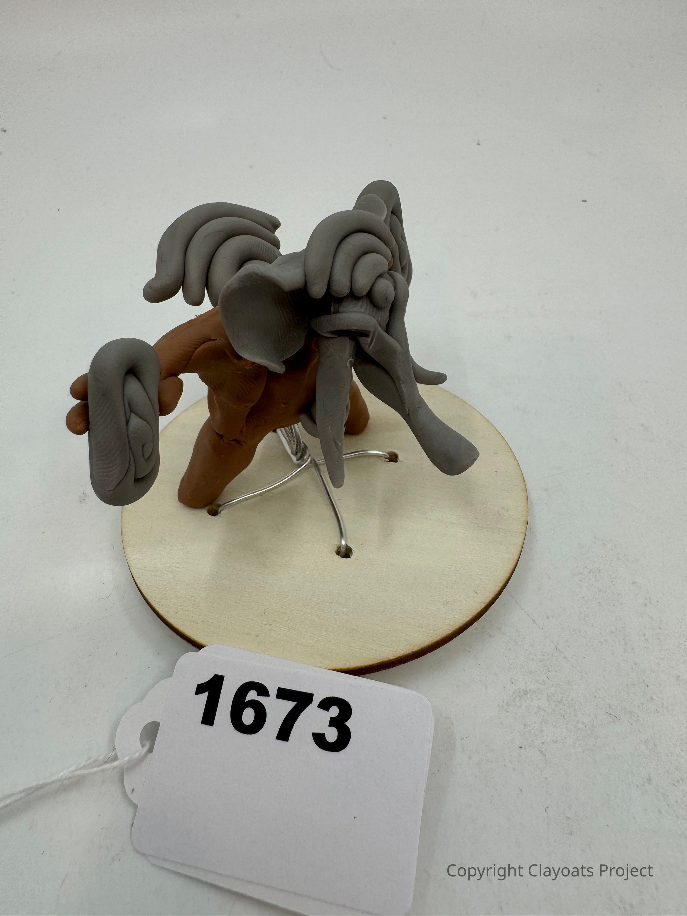 The image displays a clay sculpture resembling an abstract creature with large, flowing appendages or tentacles, perched on a simple wire stand atop a circular wooden base. This sculpture, crafted in shades of brown and grey, exhibits a sense of organic motion and creativity. The base is plain, highlighting the unique form of the creature, and in the foreground.