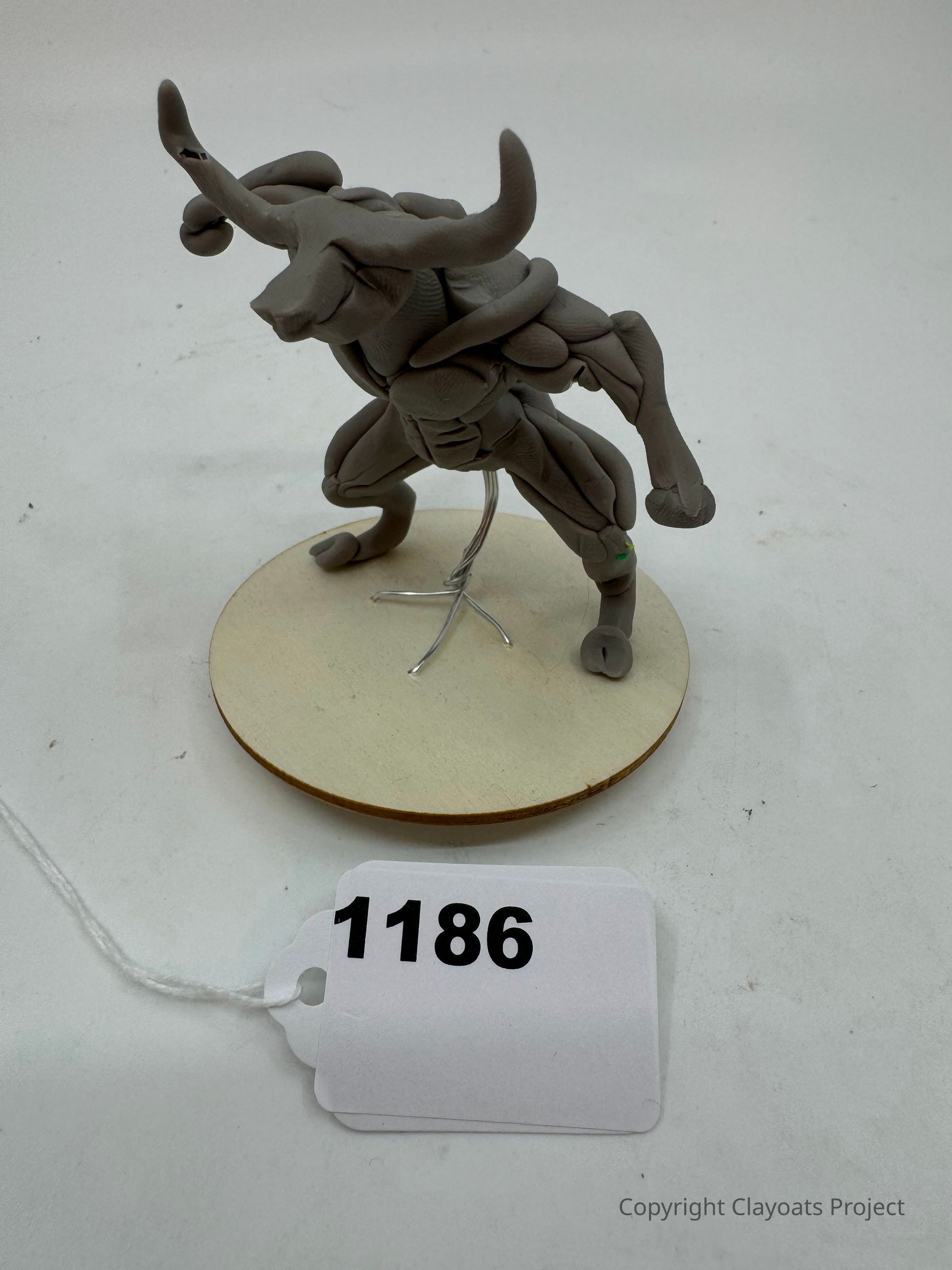 The image shows a detailed sculpture of a creature resembling a bull, crafted in a gray material and positioned on a round base. The figure is dynamic, with exaggerated muscular features and large horns, suggesting movement or an action pose. It stands on a wire support for stability. The sculpture is poised against a plain background, emphasizing the intricate detailing and artistic effort in its form.