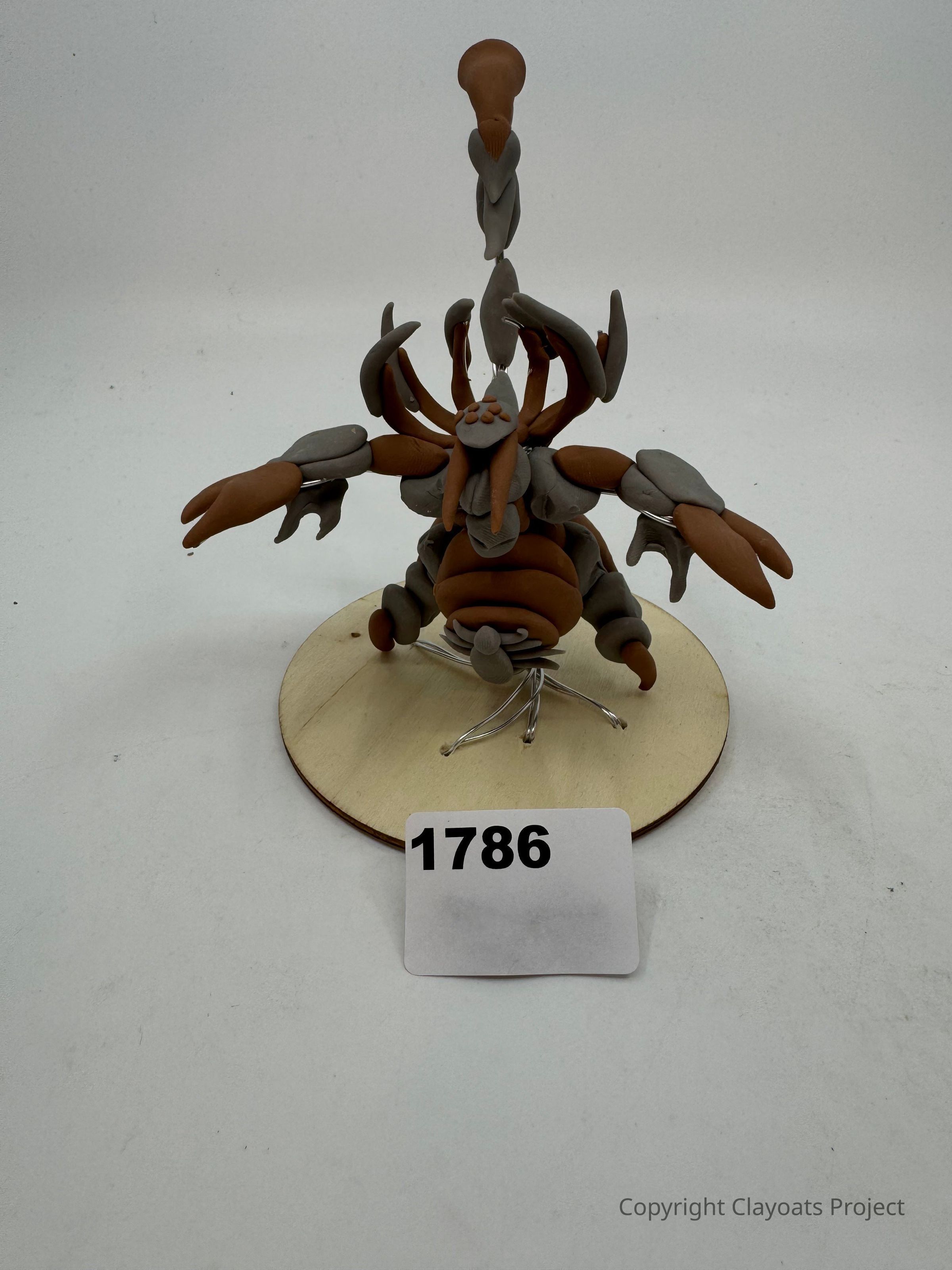 The image depicts a sculpture resembling a fantastical creature, crafted primarily from clay in shades of brown and gray, featuring multiple limbs and intricate detailing, perched on a circular wooden base. The creature's tail curves upward in a striking pose, while its claws and body parts are meticulously modeled. The background is a neutral off-whiteo, creating a simple and clean setting that draws attention to the