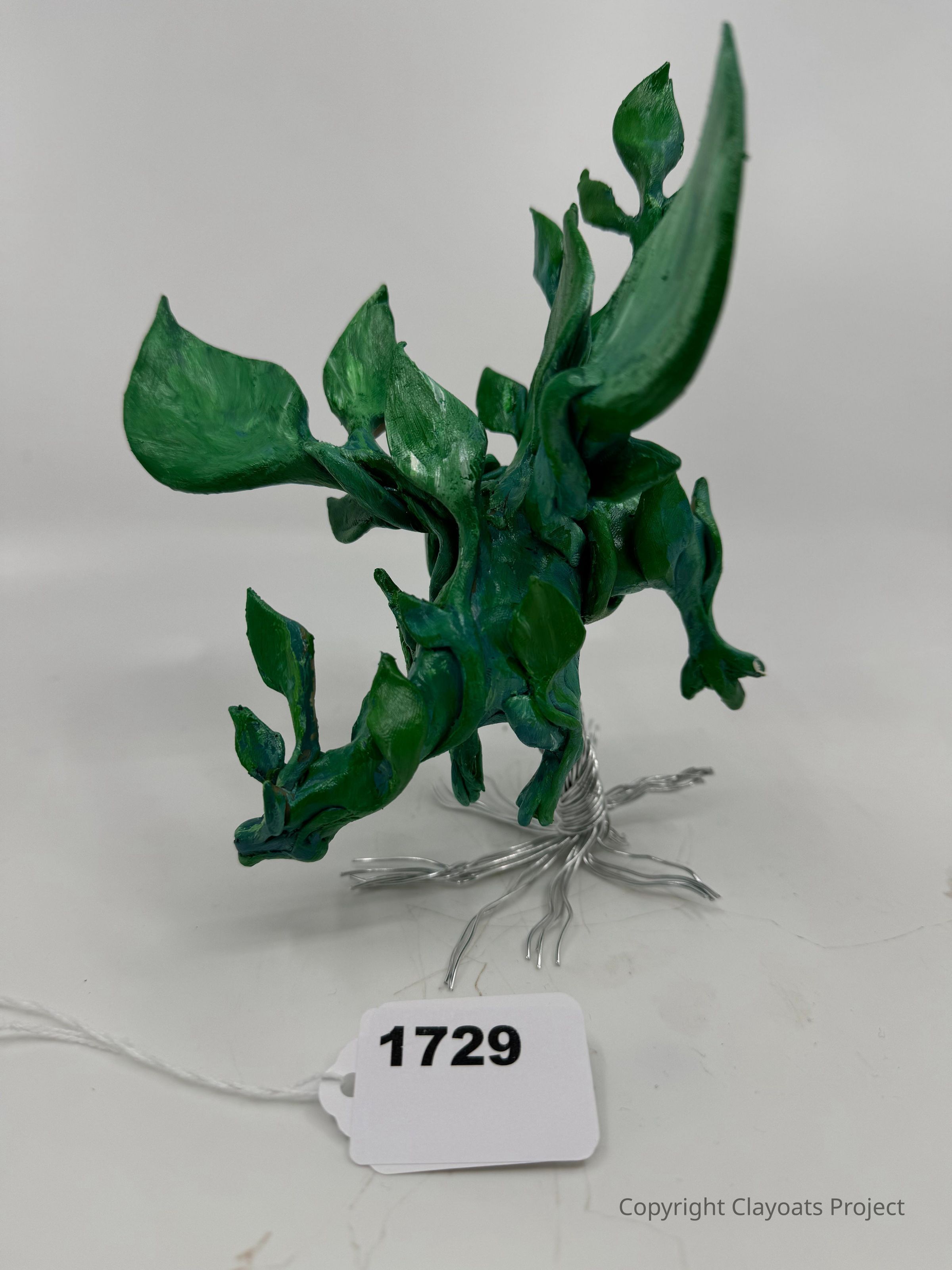 The image shows a small sculpture crafted from green material that resembles organic shapes, resembling leaves or plant-like forms, intricately arranged on a base formed by twisted silver wires that resemble roots or branches. The entire composition evokes an abstract interpretation of nature, combining artistic creativity with natural elements, set against a plain background that highlights the sculpture's intricate design and vivid color,
