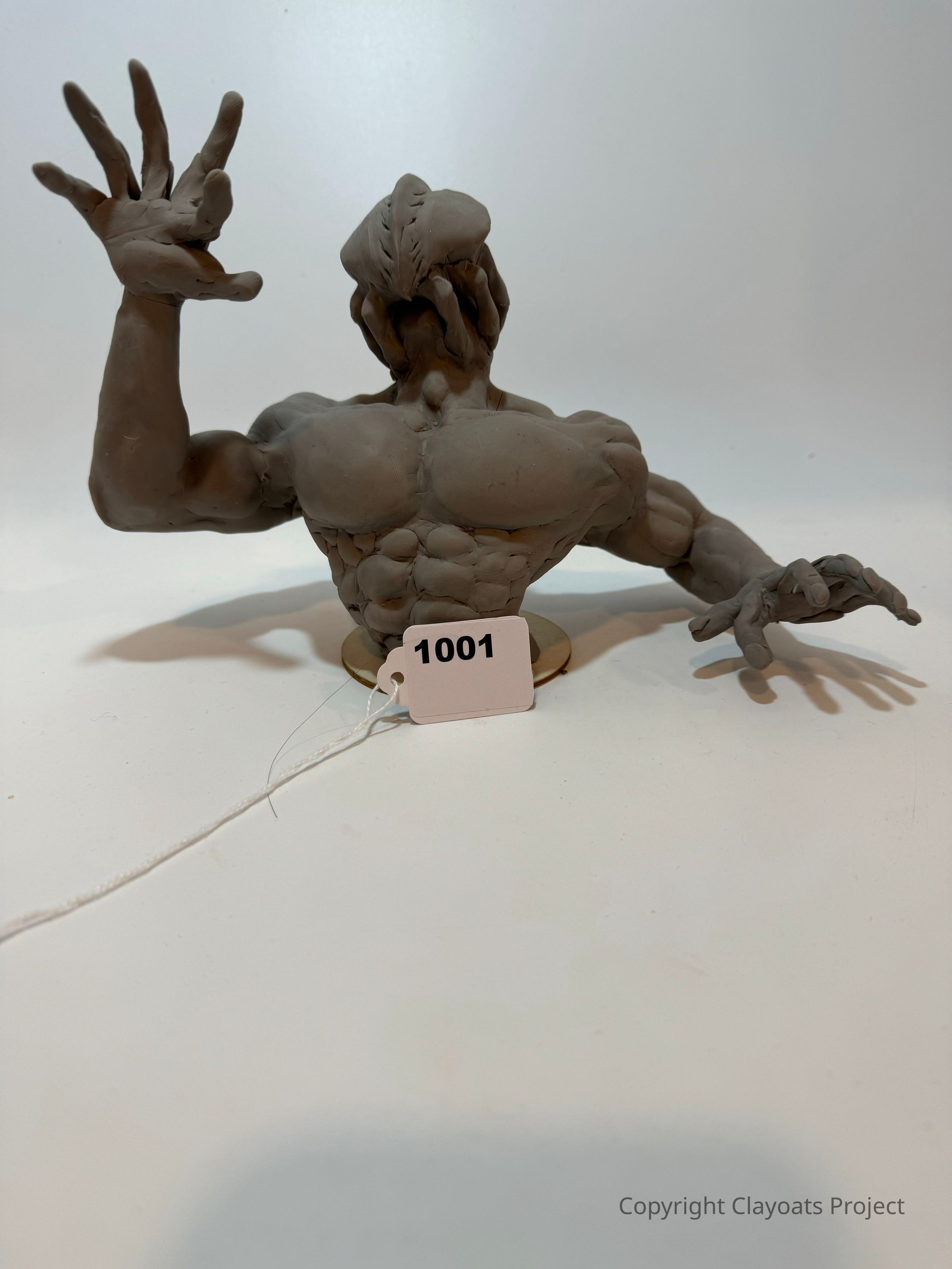 The image depicts a creatively sculpted clay model of a muscular humanoid figure with exaggerated features, including bulging muscles and a unique head structure, with one arm raised and fingers splayed, while the other is extended forward, all seated on a small base; a number tag marked "1001" is attached to the sculpture, suggesting it might be part of a display or competition, emphasizing the intricate details of its form in a well-lit, neutral space that highlights its craftsmanship and artistry.