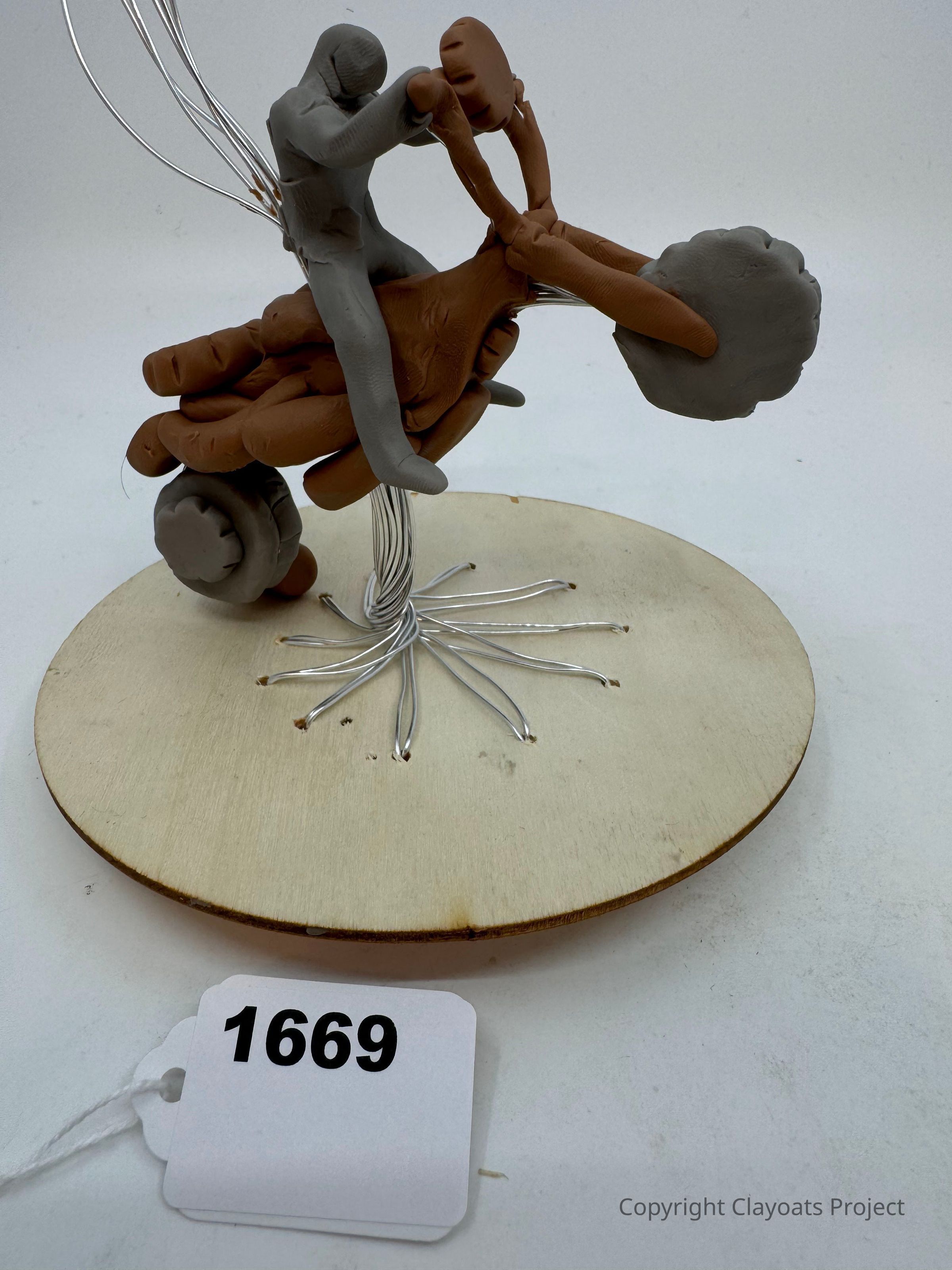 The image depicts a creative sculpture on a wooden base, featuring a gray, faceless figure riding a motorcycle made of brown clay, with silver wires extending upward from its back wheel area. The motorcycle structure has prominent wheels and exaggerated handlebars, suggesting motion and a playful approach to design. The piece sits on a round platform, with several wires spread out and attached at the bottom.