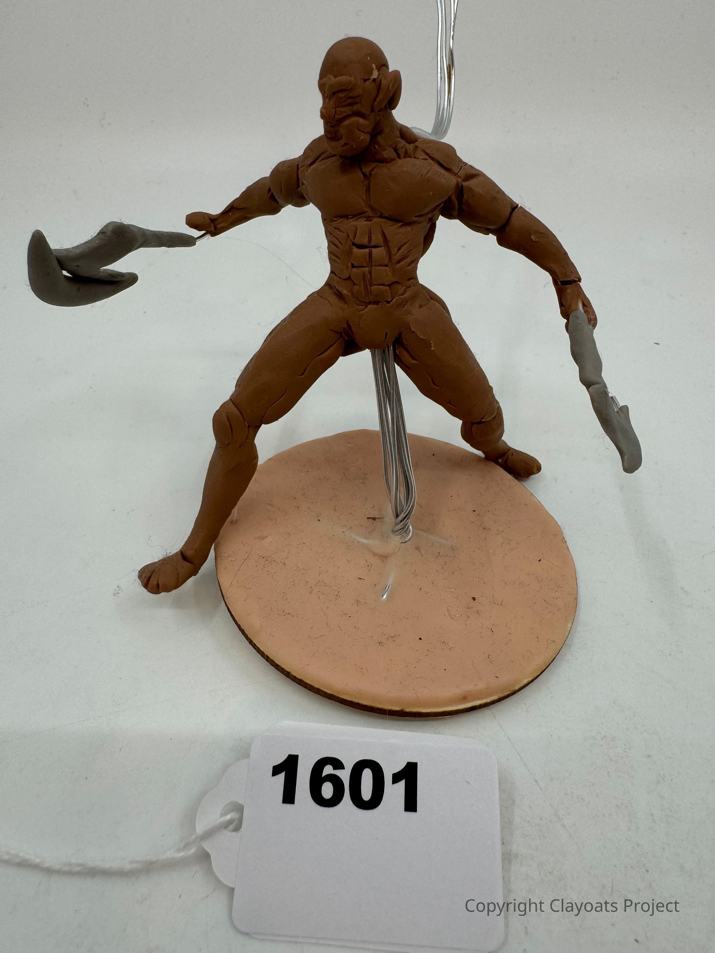 The image shows a clay sculpture of a muscular figure holding weapons in both hands, standing on a round base with a metal support rod at the back. The sculpture is positioned in a dynamic warrior pose, with detailed anatomical features. The model's brown and gray colors contribute to its distinct and striking appearance.