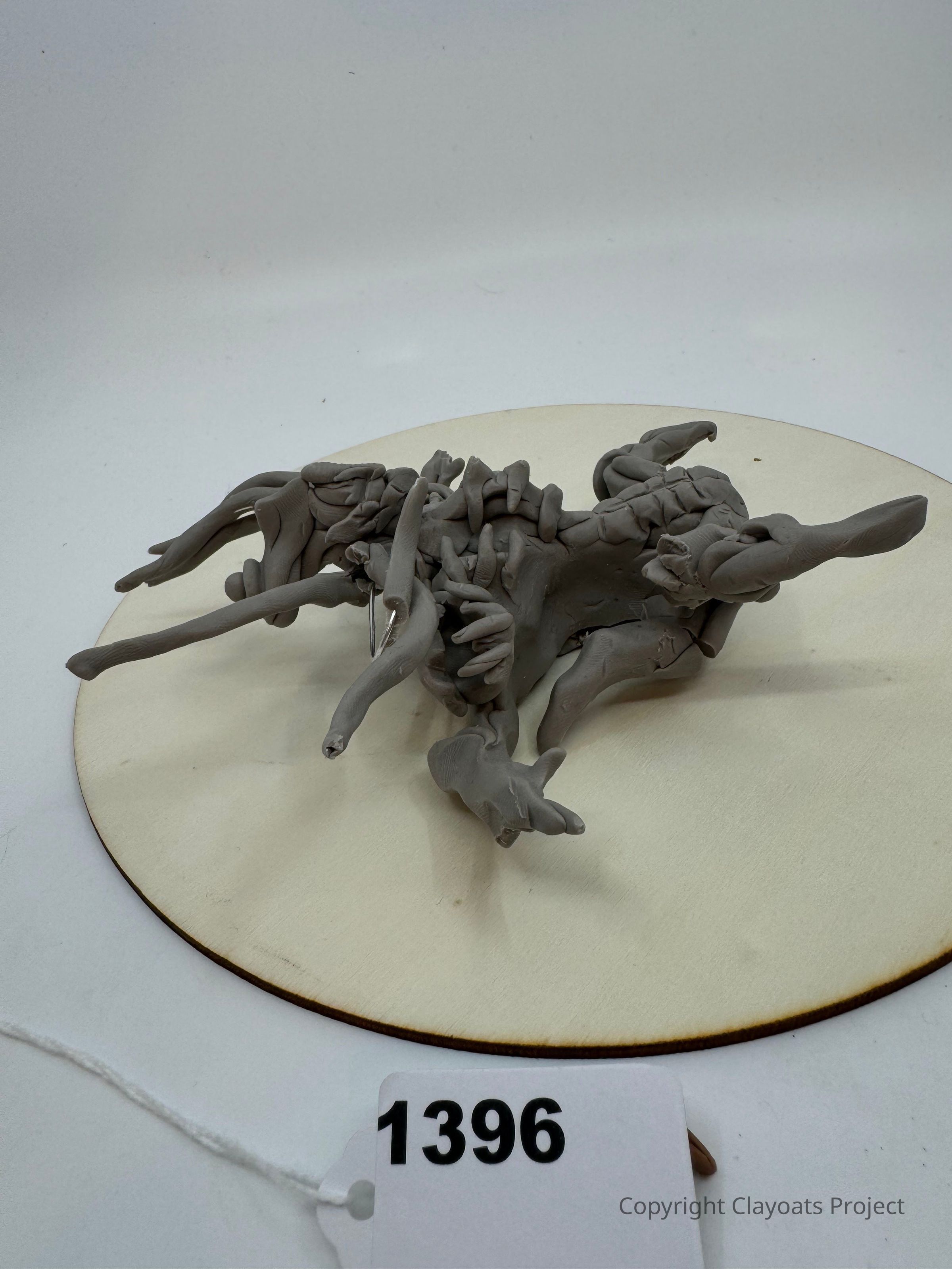 The image shows a complex, abstract sculpture made of gray clay, featuring intertwining, organic forms with elongated and twisted appendages, resting on a circular white platform. The intricate design appears dynamic, capturing a sense of motion and fluidity. The background is neutral with soft lighting that highlights the texture and shadows of the clay structure, emphasizing its artistic and