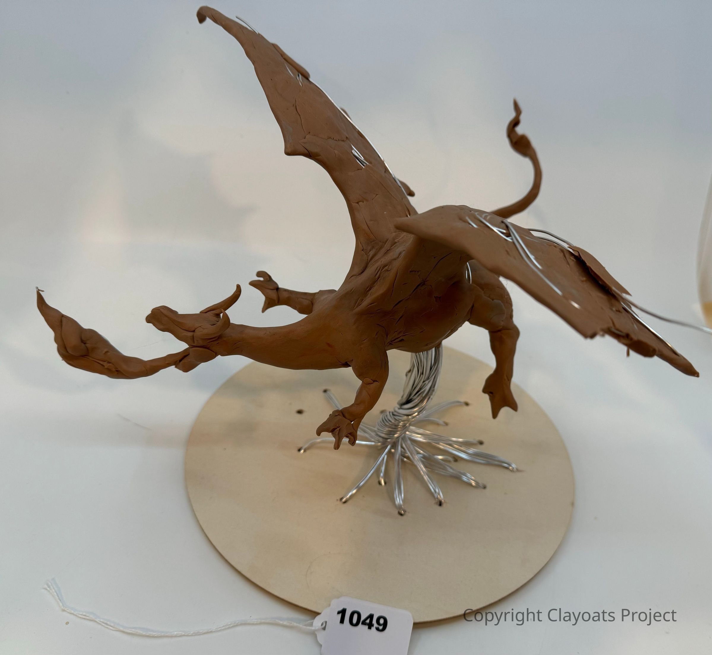 The image shows a detailed sculpture of a dragon crafted from clay, featuring a dynamic pose with its wings spread wide, tail curled upward, and an open mouth revealing a fierce expression, while the structure underneath is supported by an intricately twisted metal wire base positioned on a circular platform; emphasizing the artist's skill in capturing the mythical creature’s essence through sculpting techniques
