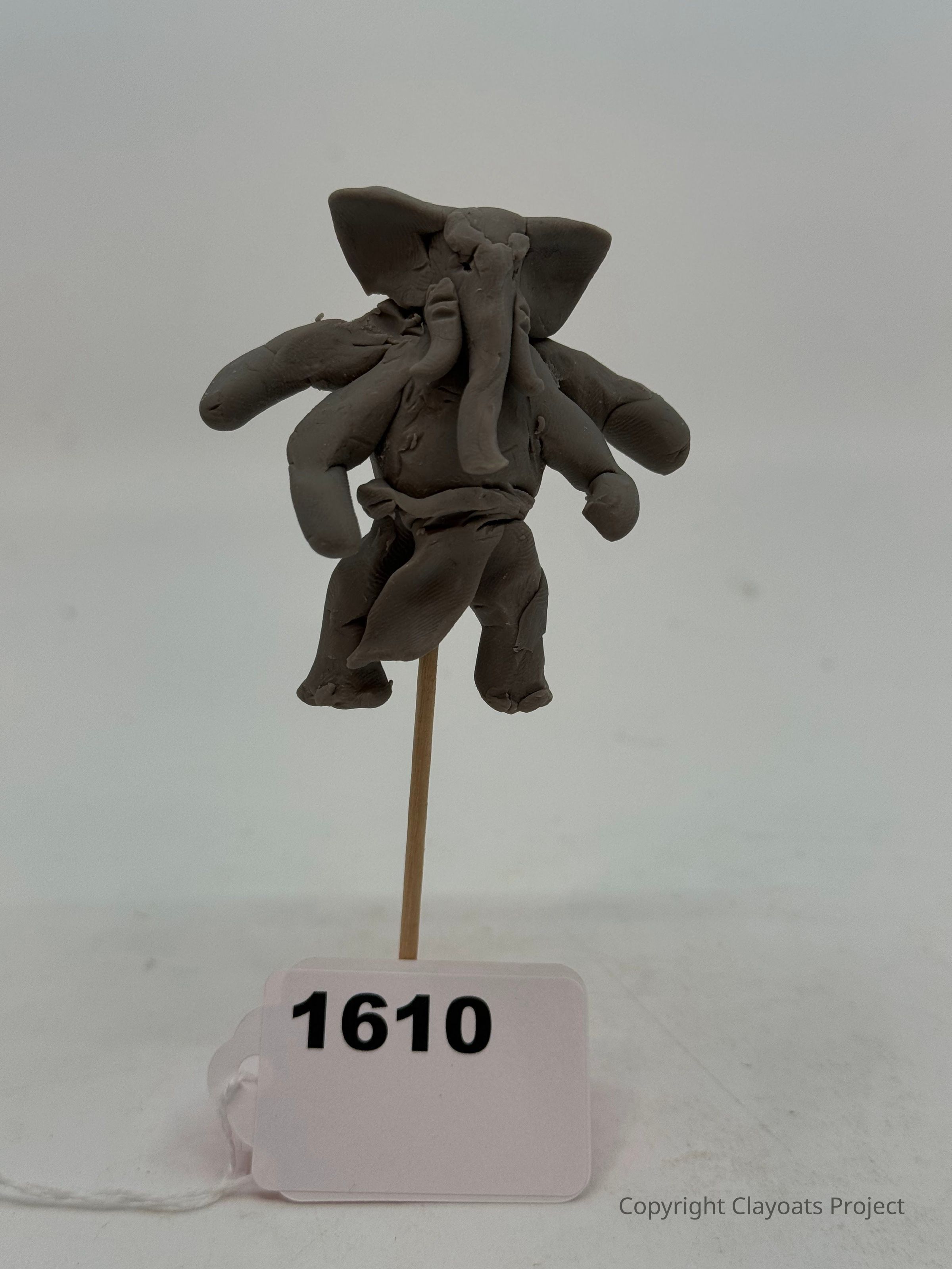 The image displays a small clay sculpture resembling an abstract elephant with multiple limbs, perched on a thin stick secured in a white base, all set against a plain light background that highlights the artistry and texture of the sculpture, which is crafted with visible detailing and a rough finish, evoking a sense of creativity and imagination while showcasing the artisan's meticulous handiwork and inventive approach to form and design, reflected in the sculpture's unusual
