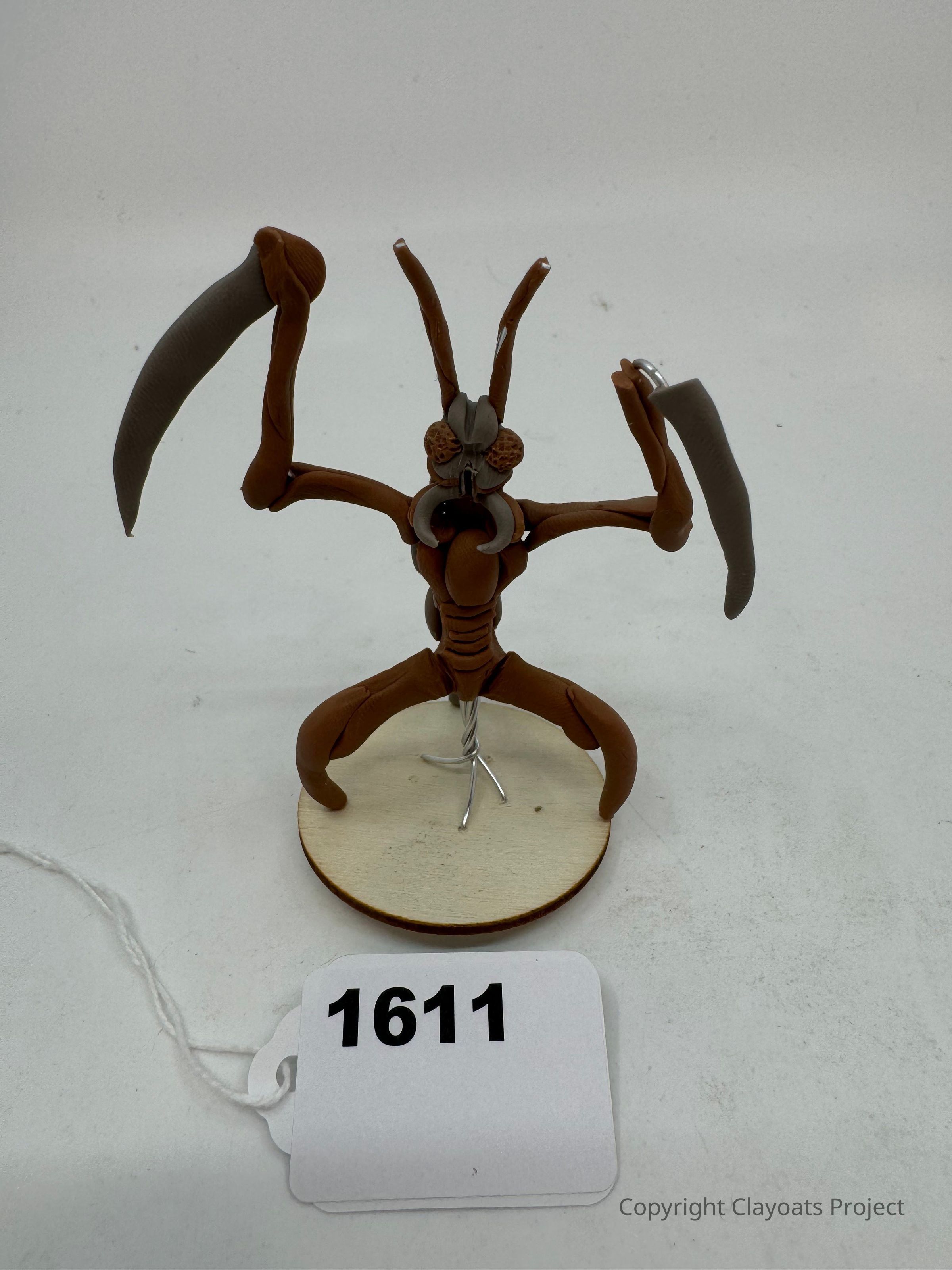 The image shows a sculpted figure resembling a praying mantis, crafted with a brown material and posed standing on a circular wooden base. The mantis has two prominent antennae and is designed with raised forelimbs, emphasizing its distinctive, scythe-like arms. The overall presentation highlights the intricate details of the insect model against a plain white background, making it the central focus of the image while showcasing