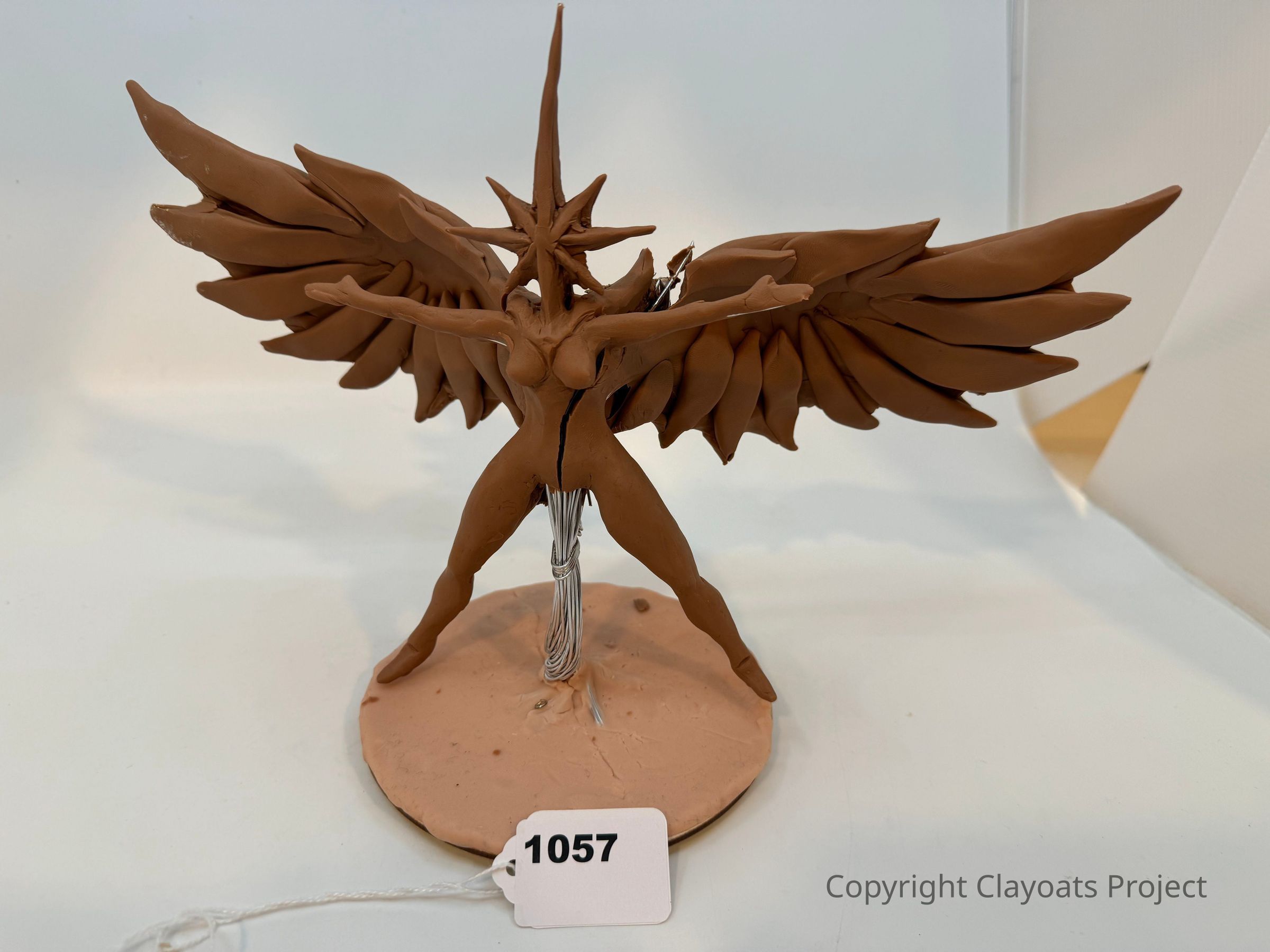 The image shows a detailed clay sculpture of an abstract winged figure, depicted with outstretched arms and large, dramatic wings extending symmetrically from its back, standing on a circular base with a protruding spiky headpiece, creating an impression of dynamic motion and balance, while a tag with the number “1057” dangles from the base, indicating perhaps a catalog or exhibit number, all set against a neutral background that highlights the intricate craftsmanship and imaginative design of the artwork.
