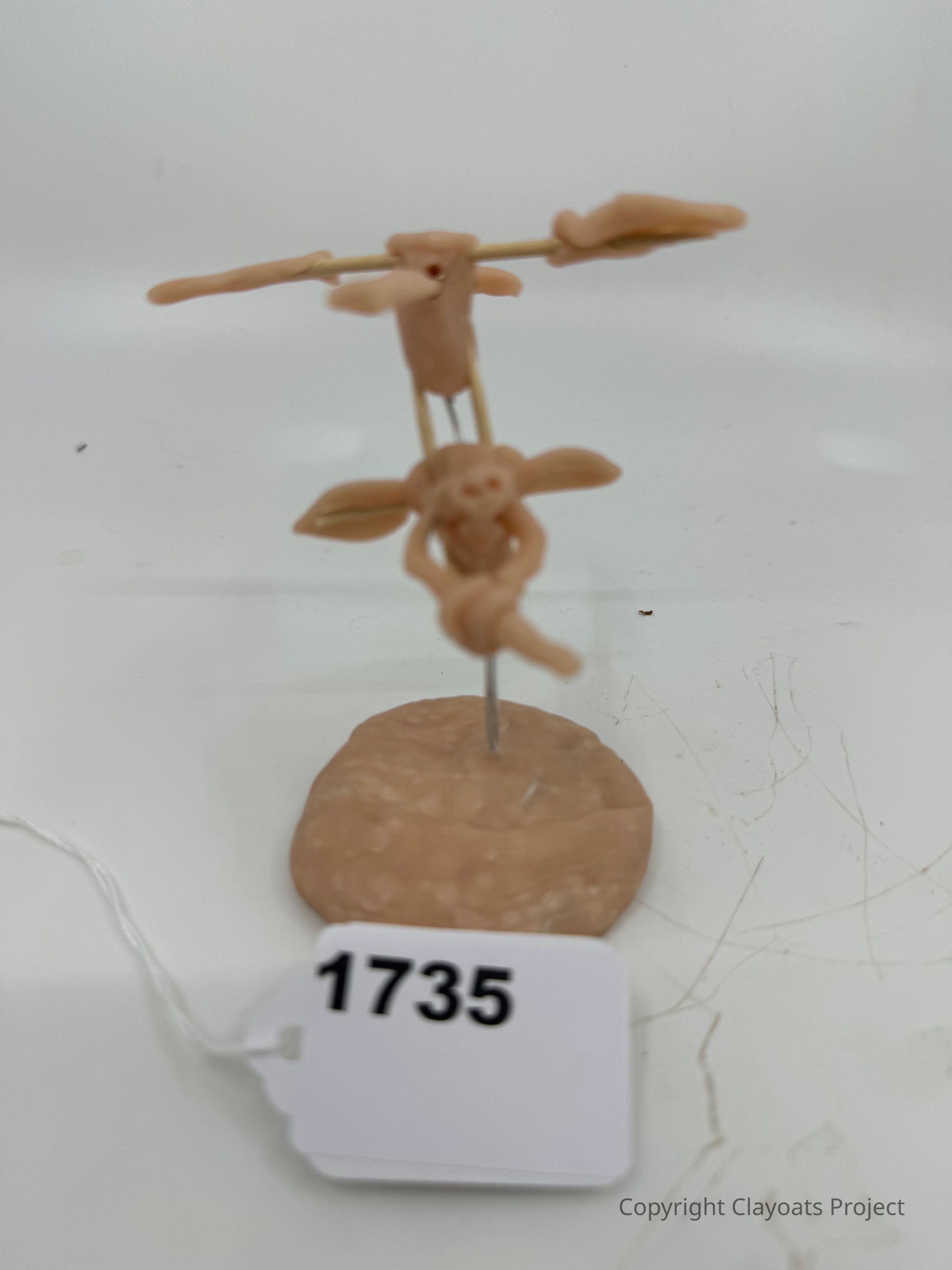 The image depicts a small clay sculpture featuring two anthropomorphic figures: one appears to be balancing on a horizontal stick with outstretched arms, while the other is positioned beneath, seemingly supporting or connected to the one above. Both figures are mounted on a thin vertical metal rod attached to a round, textured clay base.