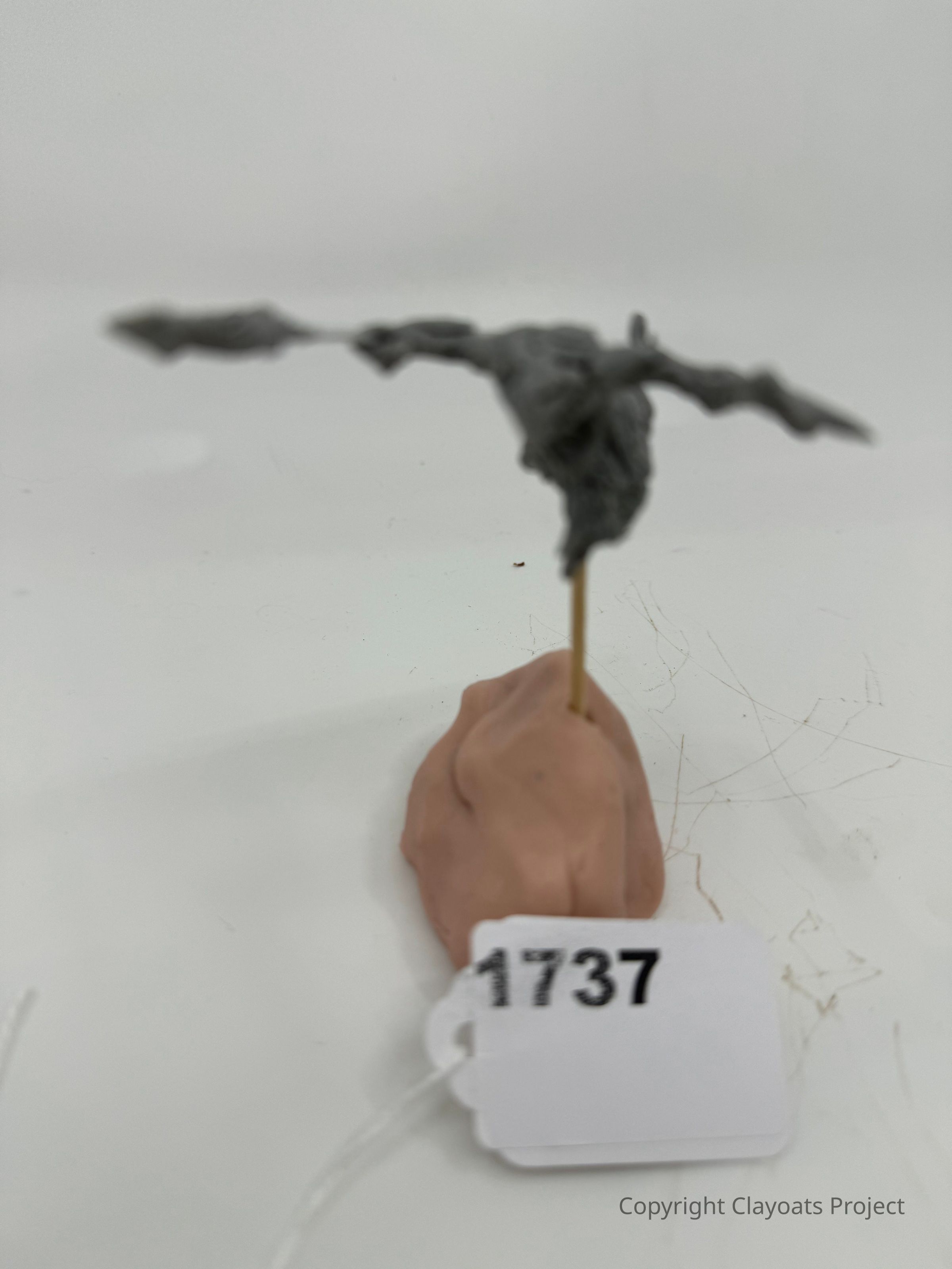 The image shows a small, abstract gray sculpture resembling a bird or bat shape, mounted on a thin wooden stick, which is inserted into a mound of clay-like material, sitting on a plain white surface. The scene appears to be photographed in a minimalist setting, with the main focus on the sculpture, which has an intriguing, rough texture and dynamic posture, suggesting motion or flight, complemented by the
