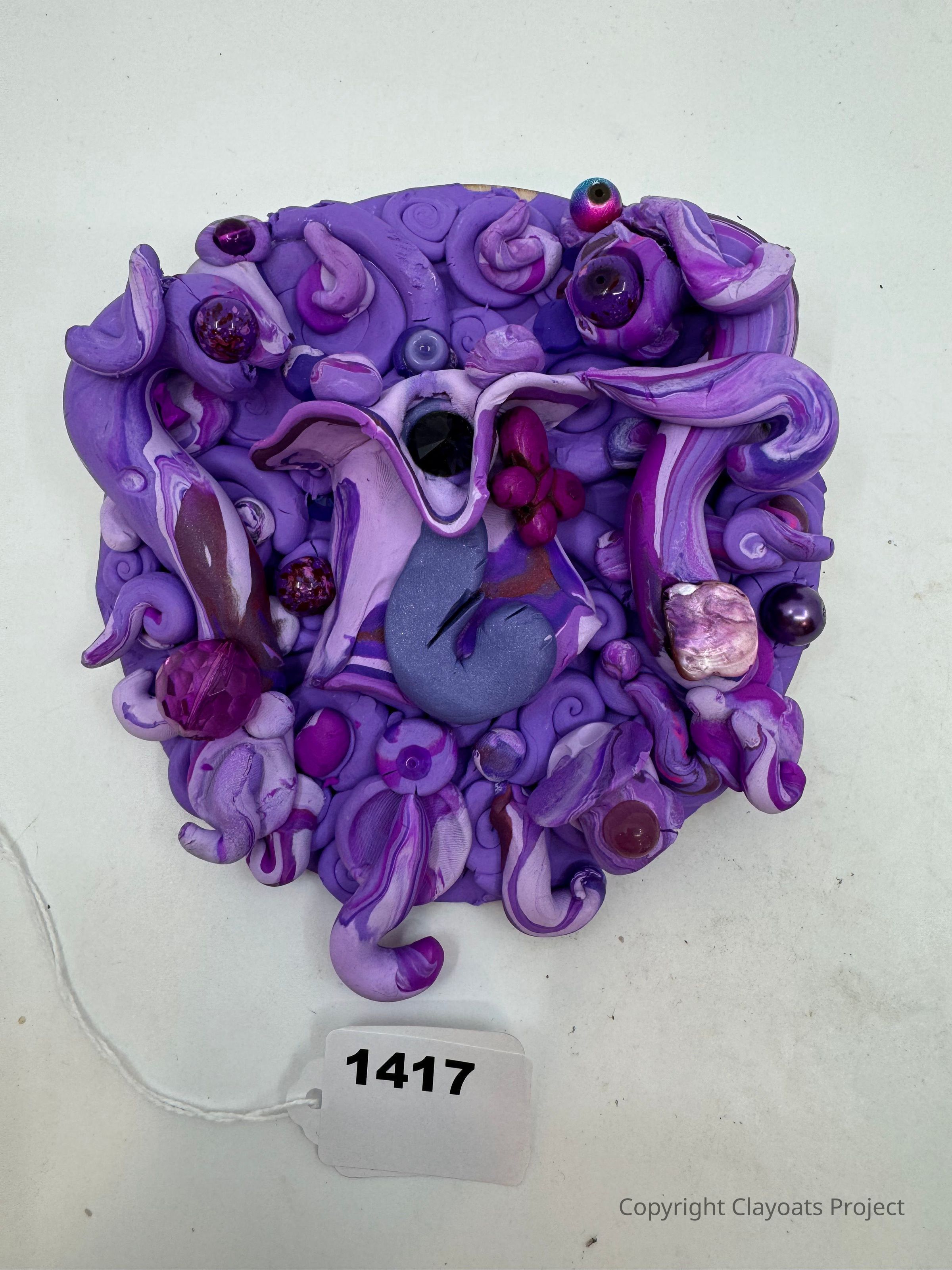 The image features an intricate, abstract sculpture composed of various swirling and twisting forms predominantly in shades of purple and violet. This three-dimensional artwork incorporates smooth and textured elements, accented by glossy, reflective spheres of different sizes. The shapes interlace and overlap, creating a sense of dynamic movement. Embedded within the sculpture are gemstones and other decorative pieces that provide visual interest and complexity to the composition. The background is a plain white, contrasting with the vivid colors and intricate details of the sculpture.