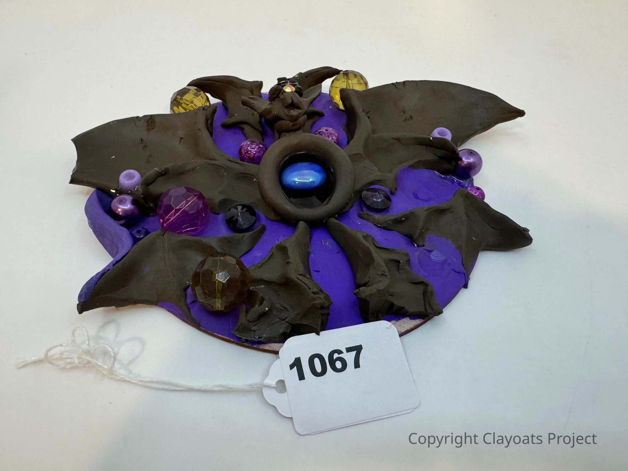 The image features a handcrafted decorative item with a purple base embellished with dark, bat-like wing shapes and adorned with an array of colorful, glossy beads and gems, including shades of purple, blue, and amber. A central, prominent dark shape seems to resemble a creature with wings, possibly a bat, surrounded by these ornamental features, creating a mesmerizing composition.