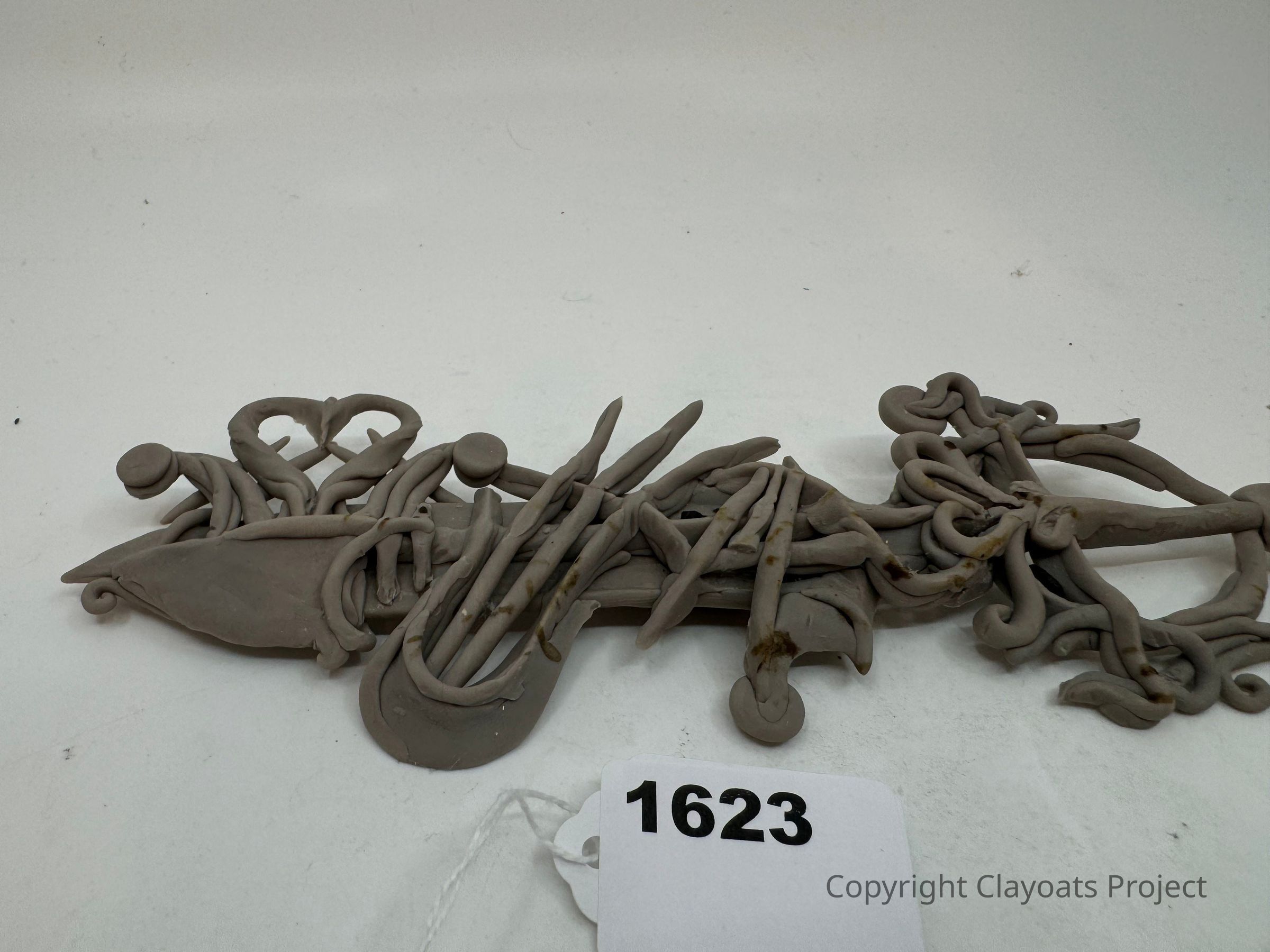 The image shows an intricate clay sculpture with an abstract, organic design featuring intertwined and looping shapes that resemble tentacles or vines, laid out on a flat surface. The piece incorporates various curves and twists, giving it a dynamic and flowing appearance. The background is a neutral shade, allowing the complex texture and form of the clay work to