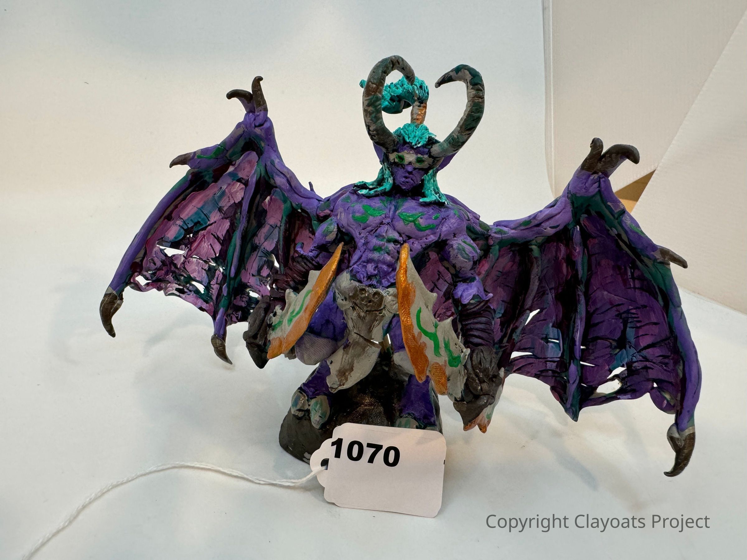 The image shows a detailed sculpture of a fantastical creature resembling a winged demon, with prominent, curved horns on its head and large, tattered wings spread wide. The creature is predominantly colored in shades of purple and blue, with intricate patterns in green and orange across its muscular body. It holds a large, ornate weapon in its right hand, which appears to be encrusted with green designs, adding to its menacing appearance. The sculpture is positioned on a plain background.