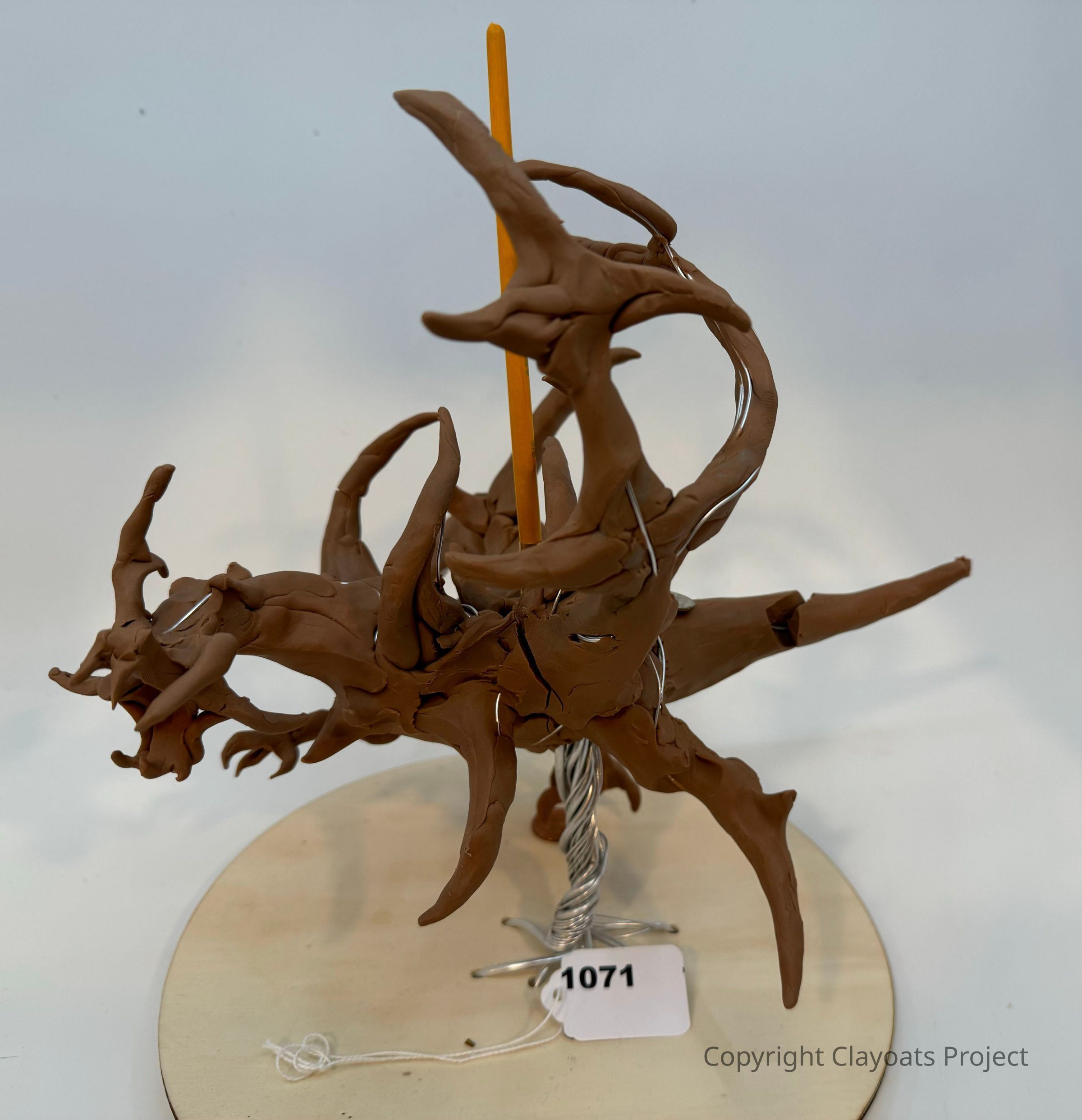 The image features an abstract clay sculpture characterized by numerous twisting and elongated spikes emanating from a central core, mounted on a wooden base. The sculpture is supported by a metal wire structure, with an orange stick extending vertically through its center. It sits on a circular base. The background is a plain white, highlighting the intricate design and earthy color of the sculpture. The overall form portrays a dynamic and organic appearance, reminiscent of tangled branches or roots.