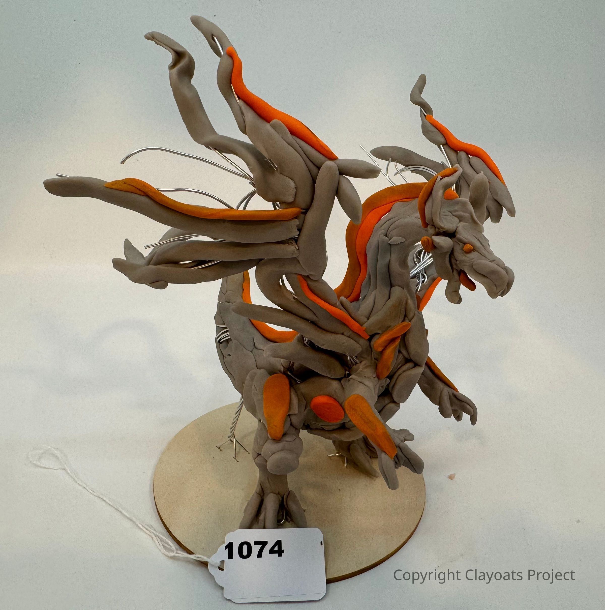 The image features a creatively sculpted dragon figurine made from a combination of clay and wire, showcasing details such as extended wings and a dynamic posture. The dragon is primarily gray with vibrant orange accents on its body, adding an eye-catching contrast to its design. It stands on a circular base, which supports its sturdy structure. The dragon's intricate details, such as its facial expression and limb positioning, highlight its fierce and mystical nature.
