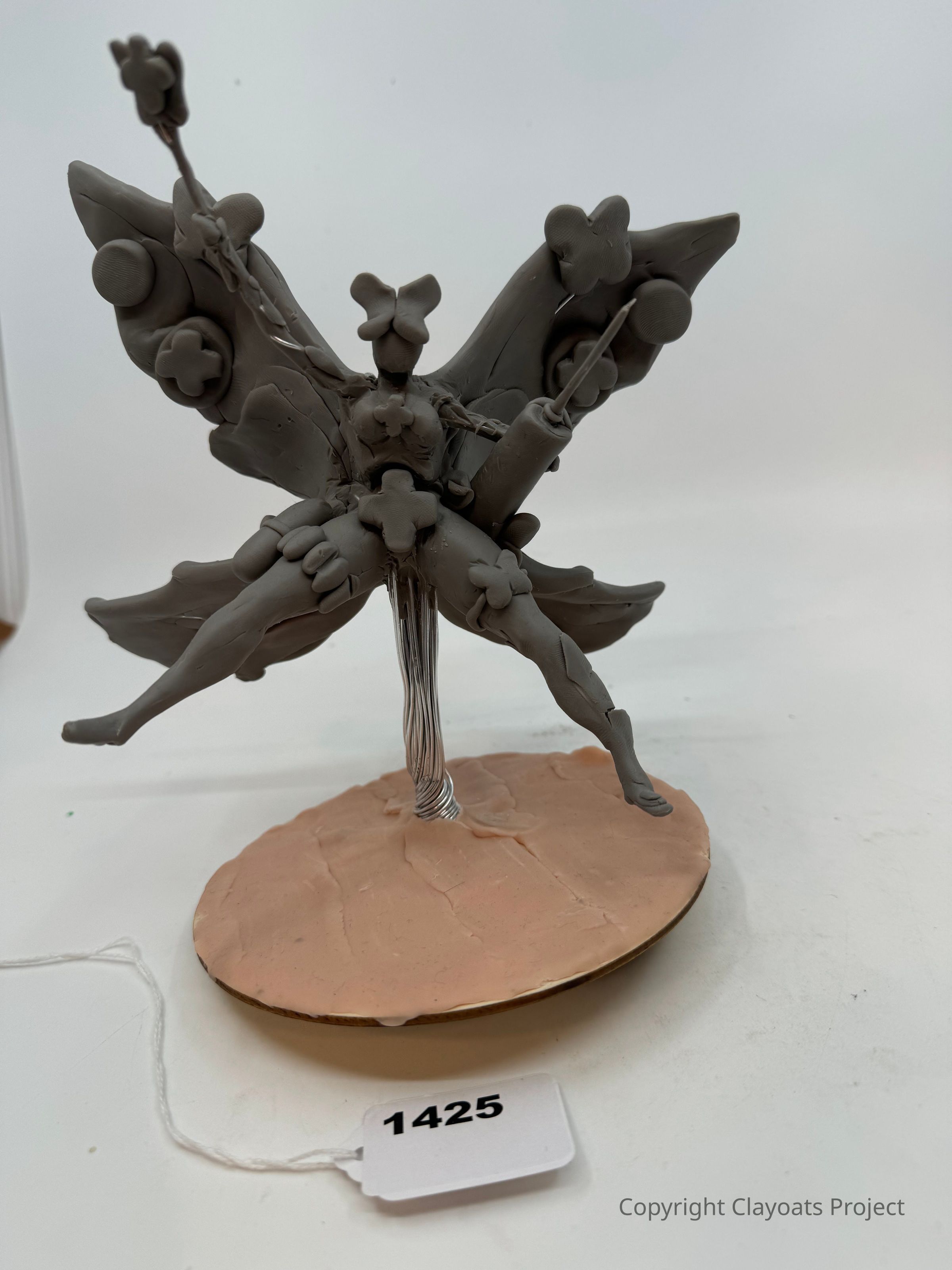 The image shows a sculptural piece made of a gray clay-like material, featuring a figure with butterfly-like wings adorned with circular and floral elements, standing gracefully on a wire attached to a textured circular base. The design is intricate, with the figure appearing dynamic and delicate, suggesting motion or flight. The base, resembling a sandy surface, complements the sculpture's theme.