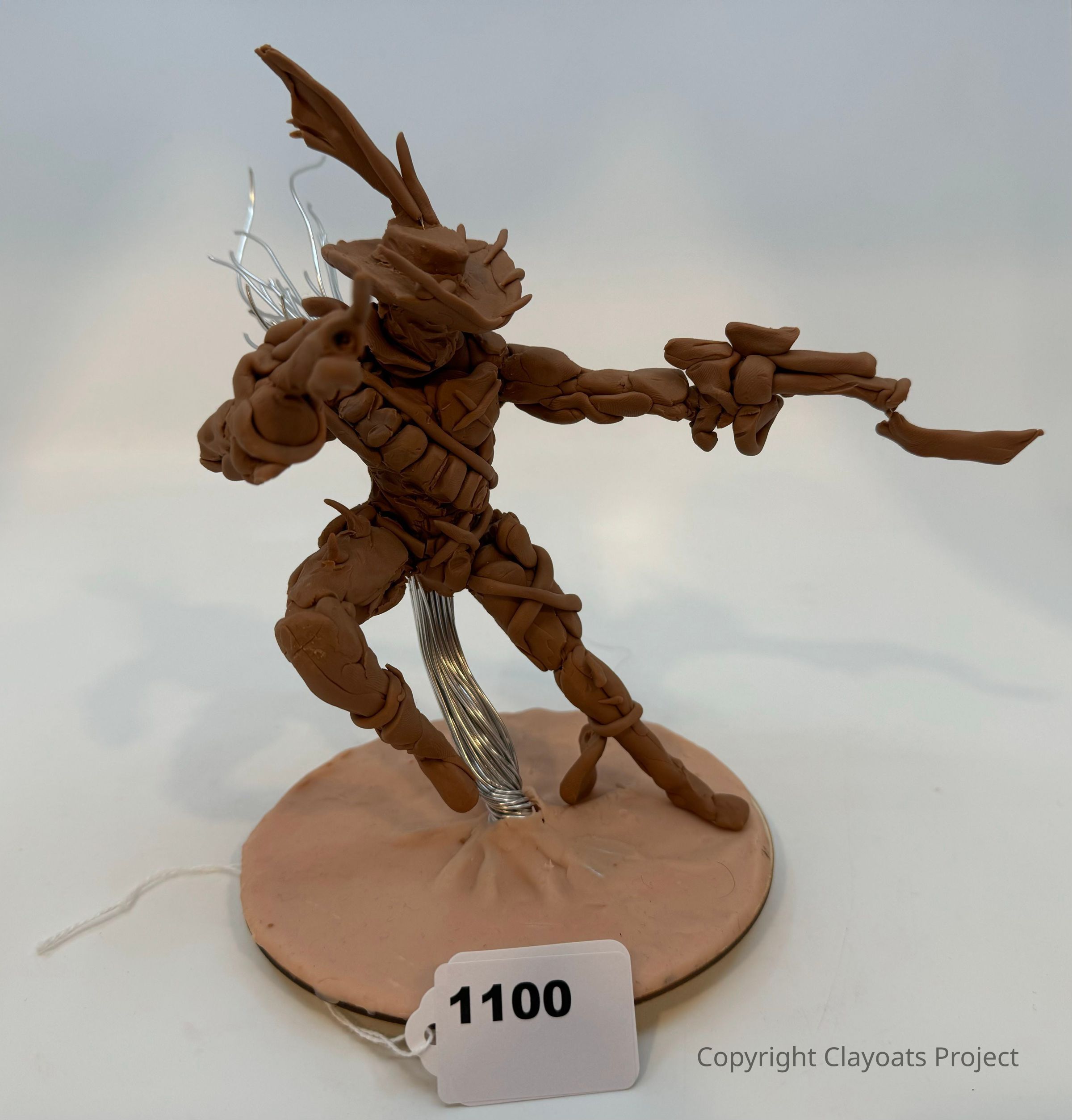 The image depicts a small, intricately modeled clay figure in an action pose on a round base, The figure appears to be a stylized humanoid character, possibly an action hero or futuristic warrior, constructed from clay with wire supports visible in the background, adding a sense of dynamic movement. The detailed craftsmanship is evident in the figure's armor-like textures and dramatic stance, suggesting an intense focus on capturing motion and expression, while the neutral background contrasts with the