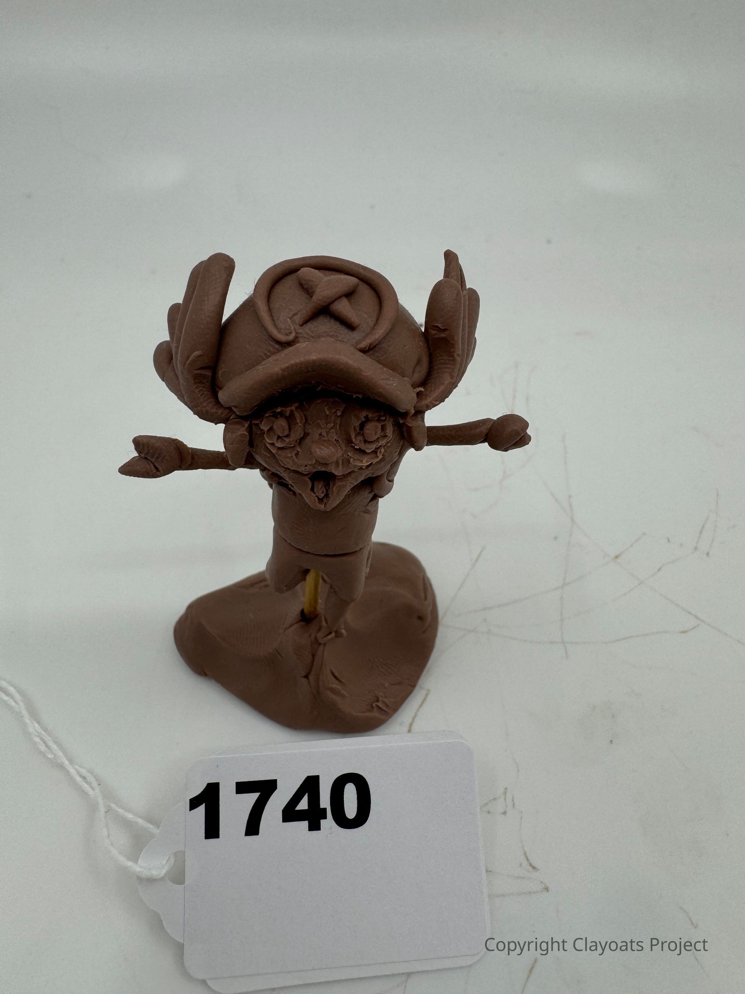 The image features a handcrafted clay model resembling a small character with a large hat, antler-like extensions, and protruding arms, standing on a base with visible texture. The hat, which has an emblem consisting of a circle and a cross inside it, sits prominently on the figure's head. The model appears creatively sculpted with detailed facial features and limbs.