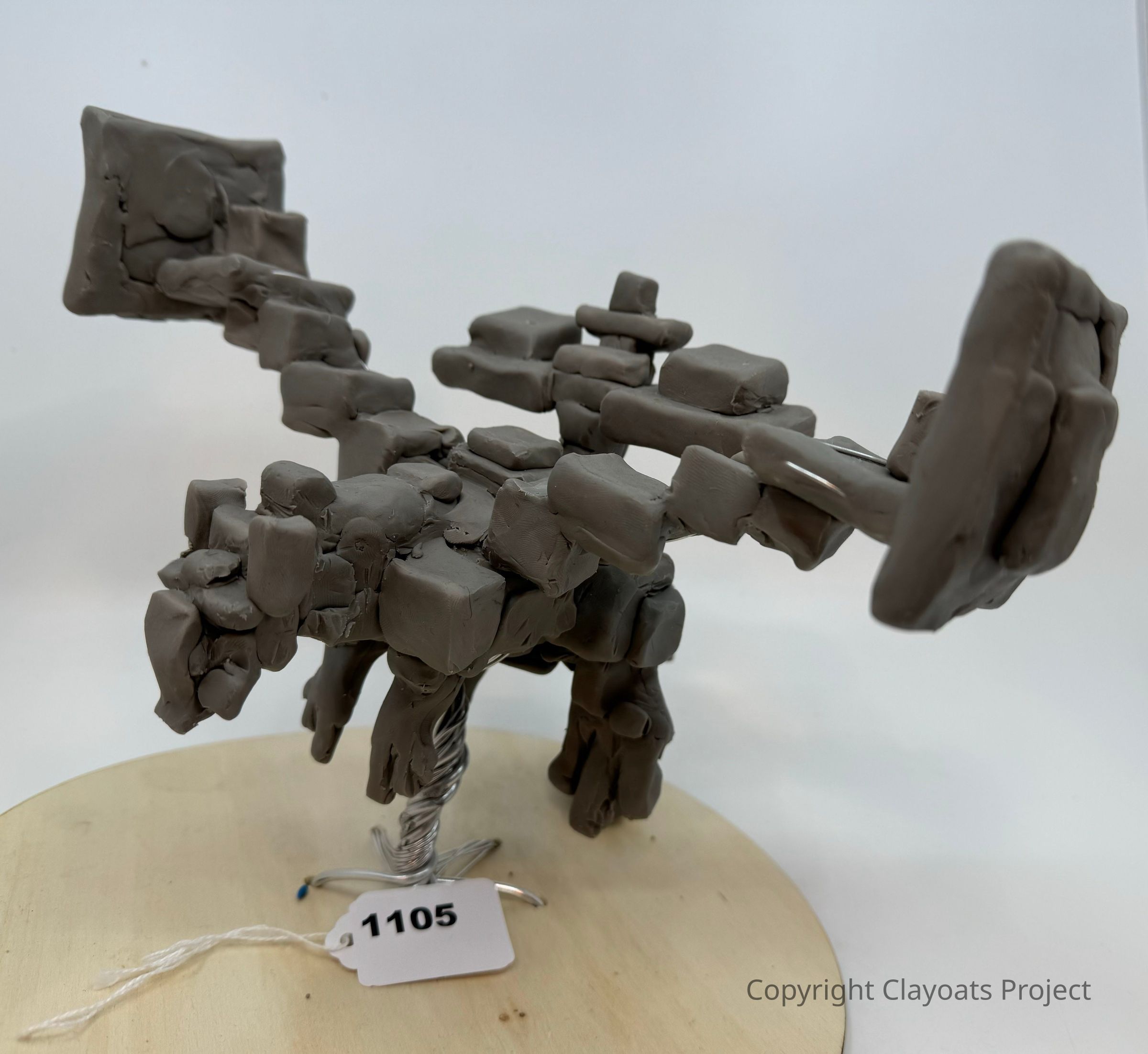 The image depicts a unique sculpture composed of numerous gray, rectangular clay blocks intricately arranged to form what appears to be an abstract animal figure, possibly resembling a dinosaur or another quadruped, standing on a circular base with a twisted wire support; while the smooth, light background helps highlight the geometric complexity and creative expression of the sculpture itself.