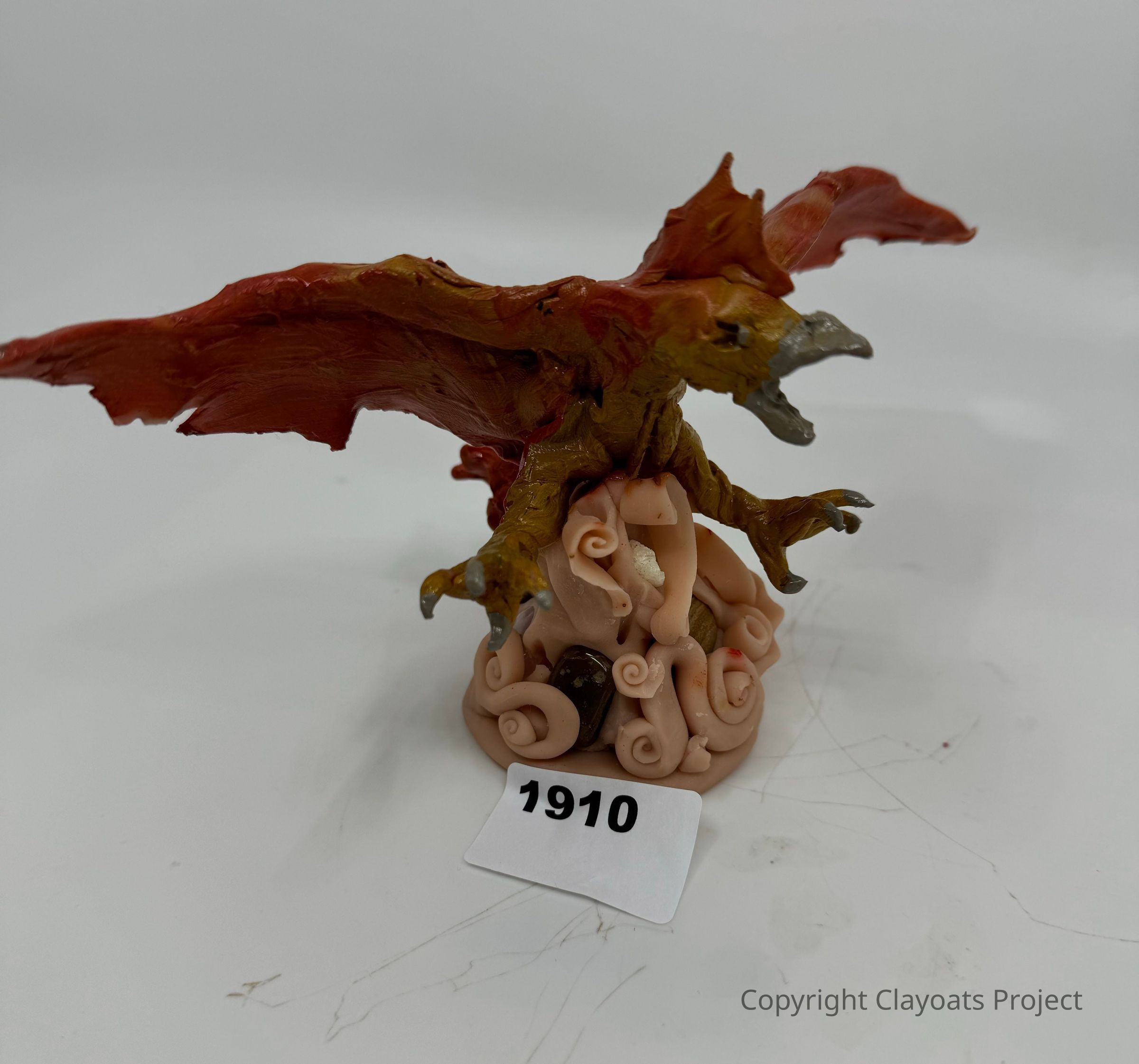 The image depicts a detailed sculpture of a fantastical creature resembling a dragon, with outstretched wings and an open mouth, perched atop a base comprised of swirled and twisted pale forms that resemble abstracted tentacles, all placed on a light surface.The dragon's body is rendered in shades of reddish-brown, with sharp, pointed claws and a textured