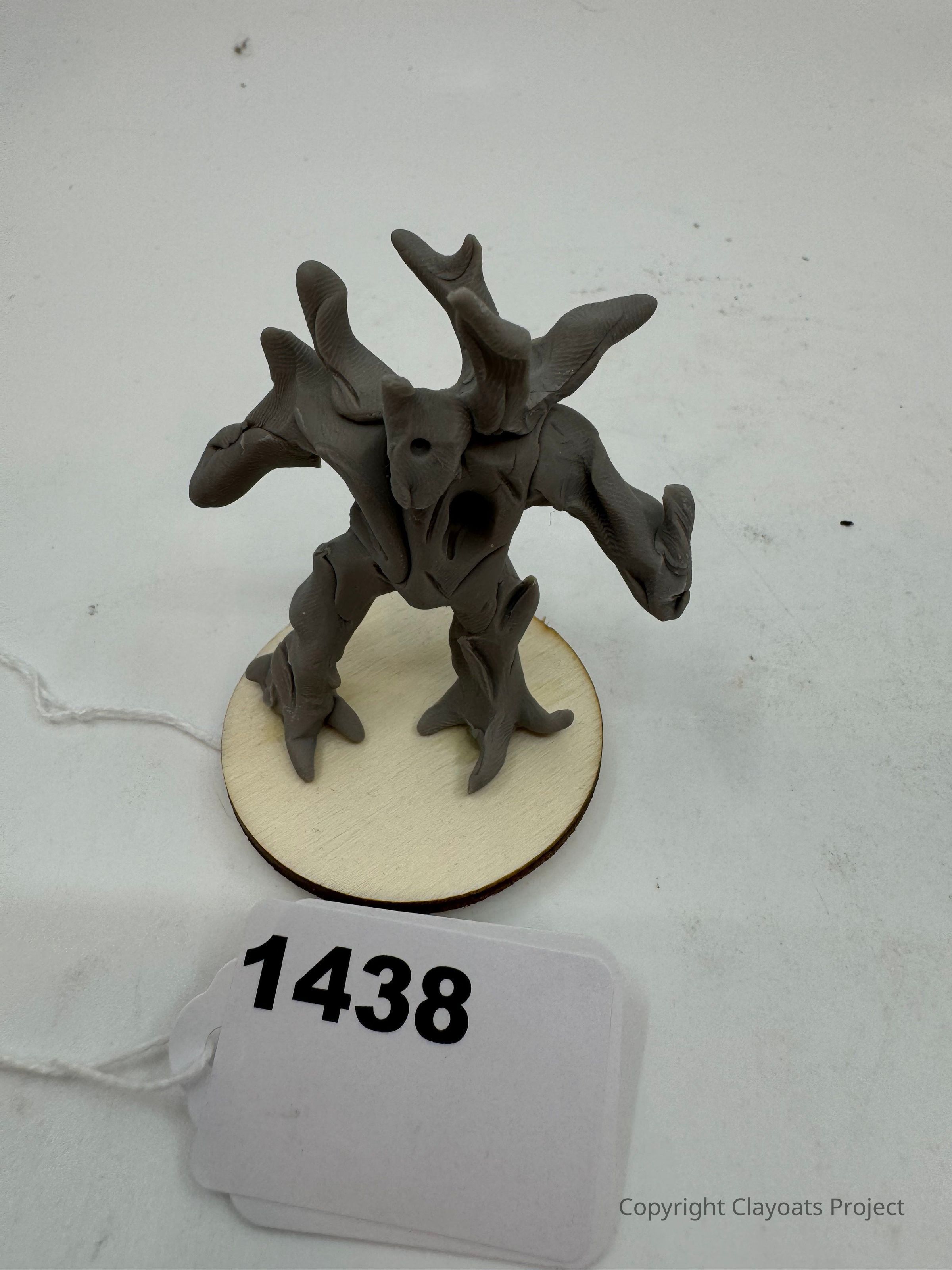 The image shows a small, intricately designed sculpture resembling a fantastical tree creature, featuring multiple twisting branches and limbs, standing on a circular wooden base. The sculpture is colored in a muted gray tone and appears to be crafted from clay or a similar material, showcasing detailed textures that mimic the rough surface of bark.