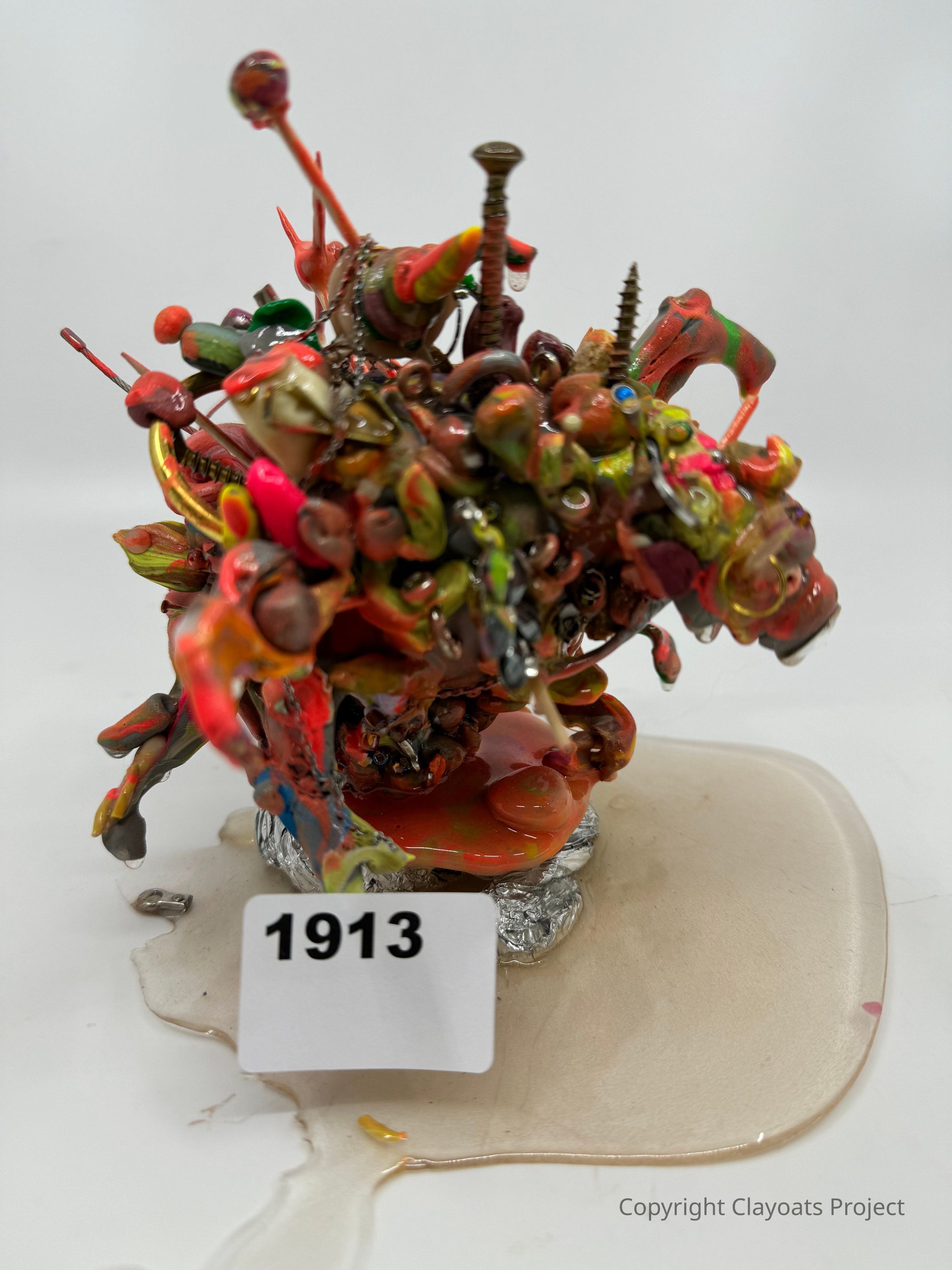 The image features a colorful, abstract sculpture made from various materials, including screws and what appears to be paint or melted plastic, creating a chaotic yet intriguing form. Its vibrant colors include red, orange, green, and yellow, with some elements protruding outward. The sculpture is placed on a surface with a glossy puddle surrounding it.