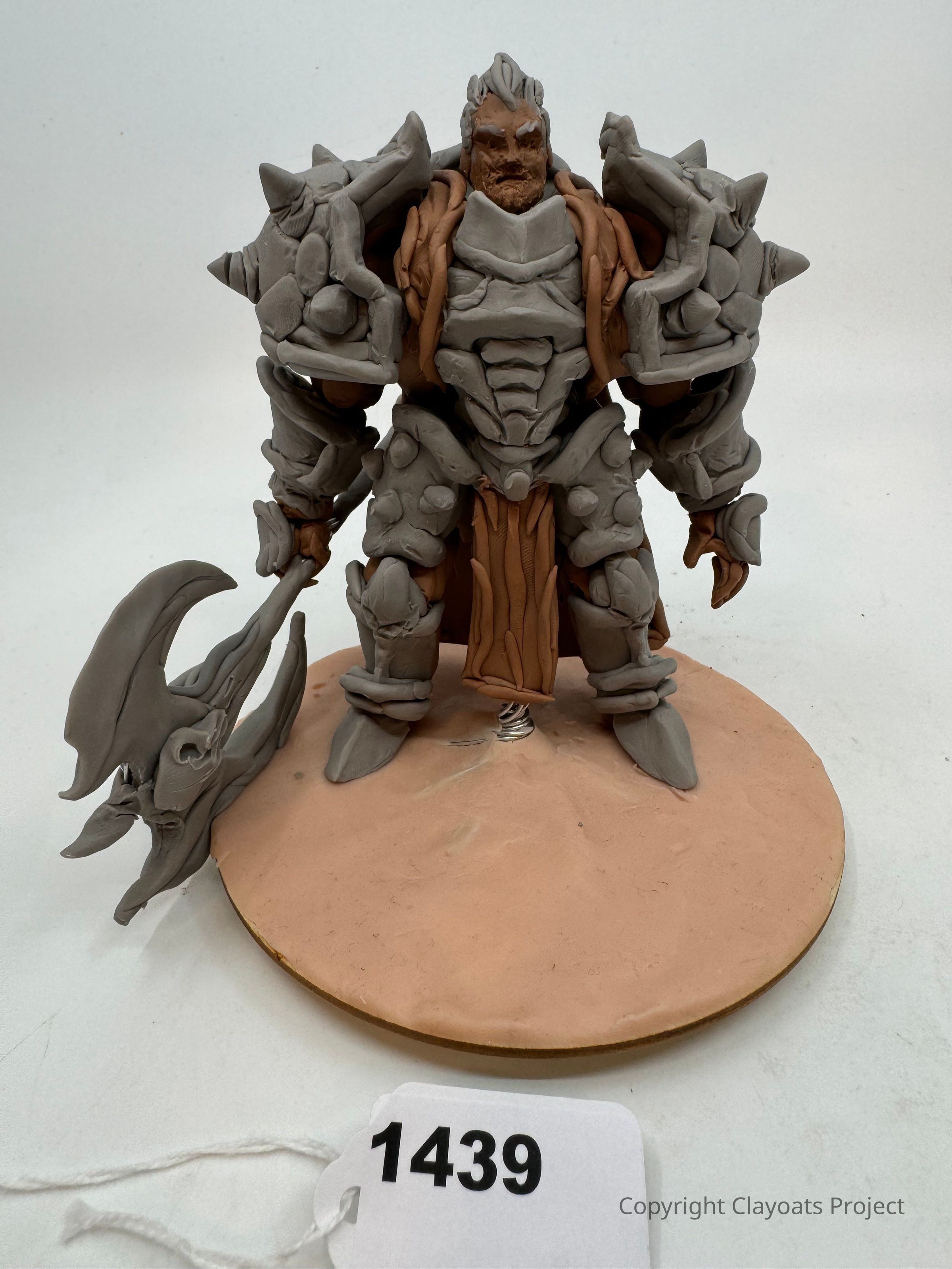 The image shows a detailed sculpture of a warrior figure standing on a round base, crafted with a mix of gray and brown materials. The warrior is heavily armored with prominent spikes on the shoulders and holds a large, axe-like weapon in the right hand. The intricate armor features various textures and details, enhancing the imposing appearance. The warrior's expression is stern, with a full beard and defined facial features. The base looks textured, resembling sand or dirt.