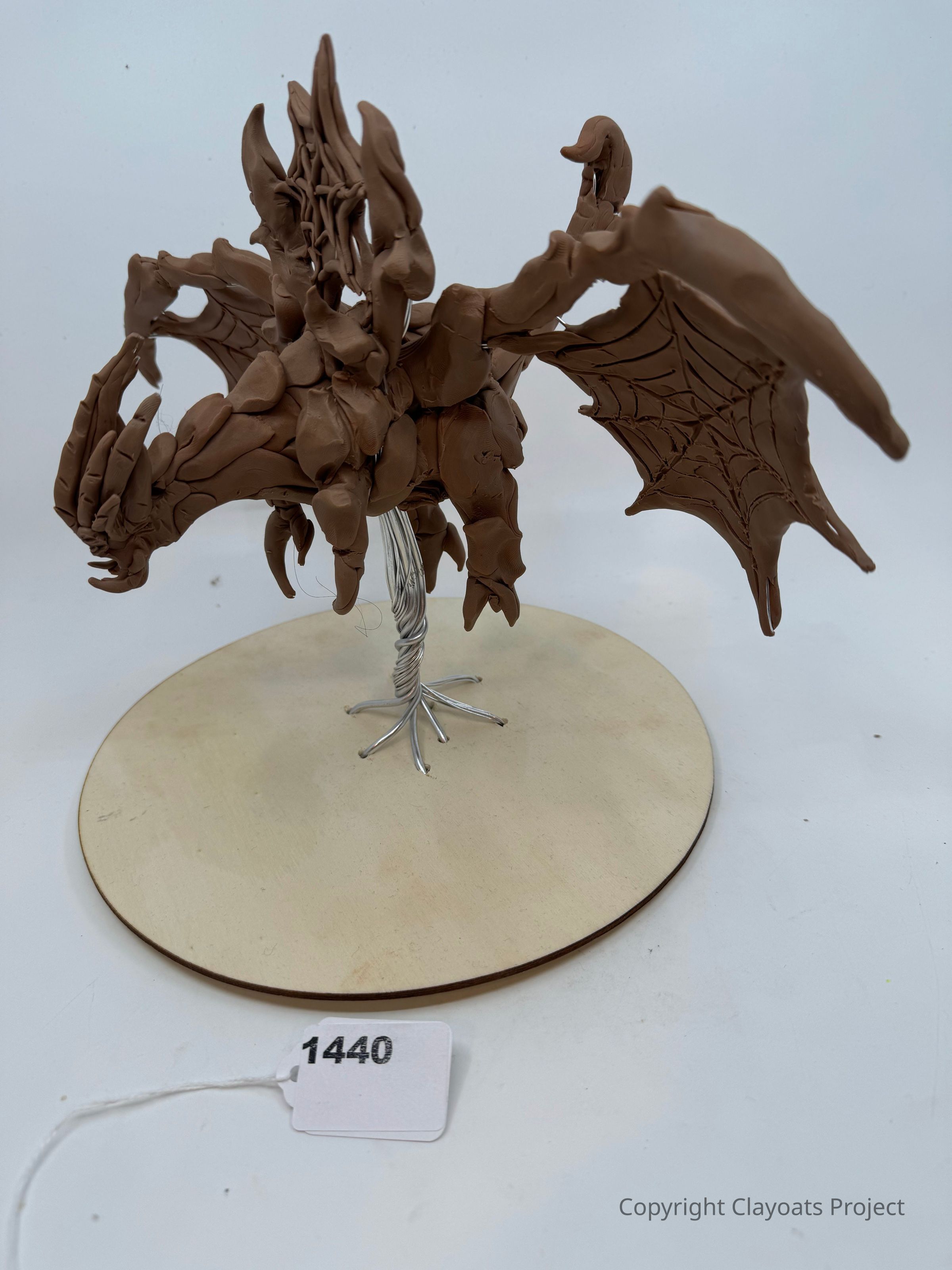 The image depicts a detailed sculpture of a fantastical dragon made from brown clay, featuring large, intricately formed wings with web-like details, and a body with exaggerated, jagged textures. The creature stands on a circular wooden base supported by a metal stem that resembles tree roots, securing the figure in place. The model is displayed against a plain white background, accentuating the craftsmanship of the dragon's fierce and complex design.