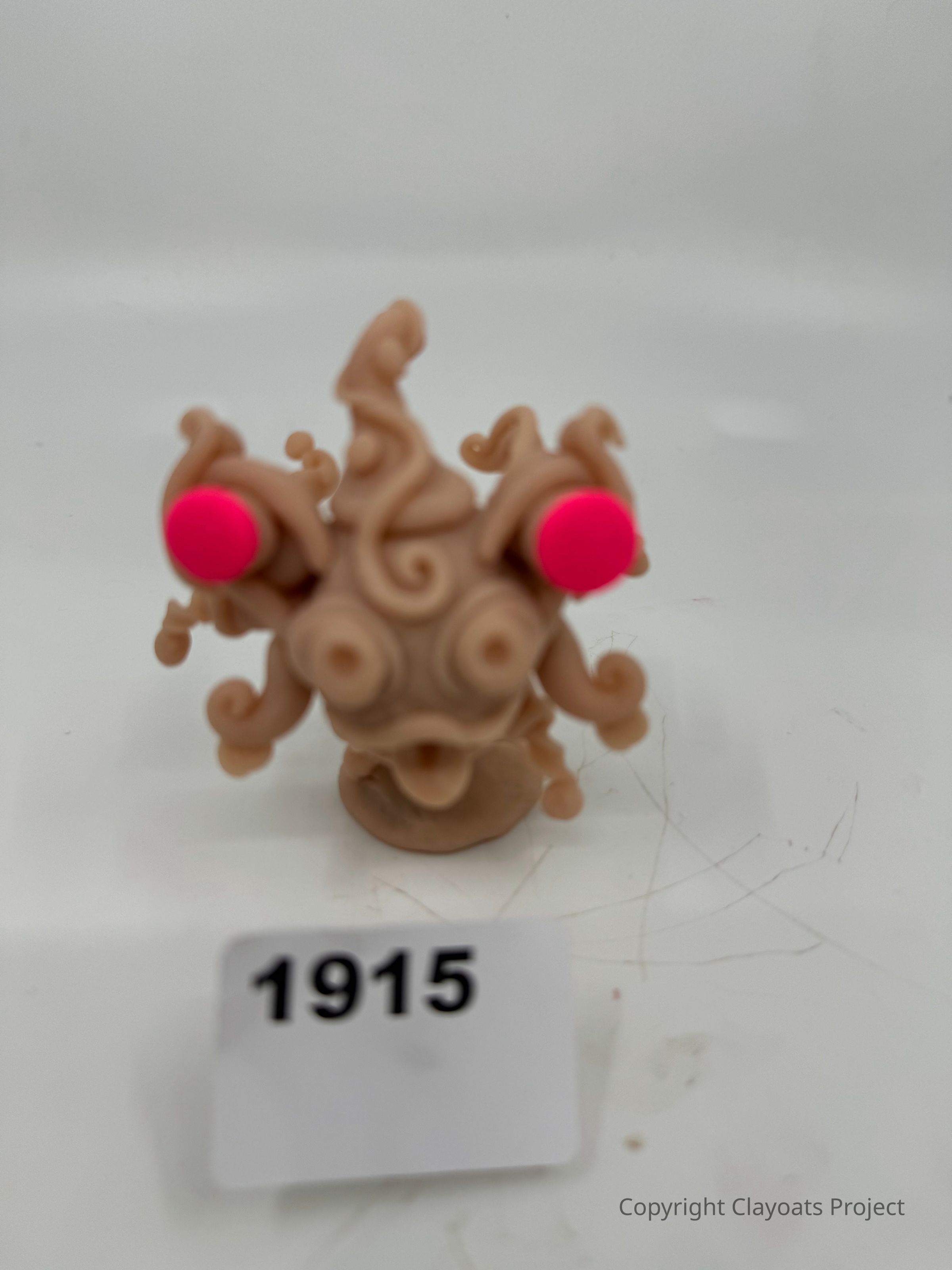 The image shows a small, abstract sculptural figure made of a beige material with an intricate design featuring swirling, tentacle-like appendages. The figure is adorned with two prominent, circular, bright pink accents resembling eyes. It stands on a plain surface, The background is a simple, unobtrusive light gray, which helps to draw attention to the peculiar and artistic