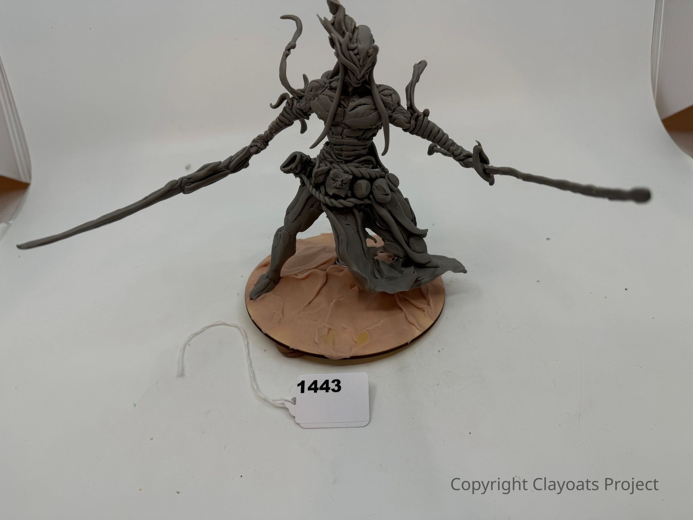 The image shows a detailed sculpture of a fantastical warrior figure standing on a round base, wielding two long swords. The character, with intricate armor and flowing garments, appears poised for action with a dynamic stance. The surface texture suggests a mix of smooth and rough areas, contributing to the detailed craftsmanship. The sculptor has likely focused on capturing motion and character with curled elements on the helmet and flowing attire.