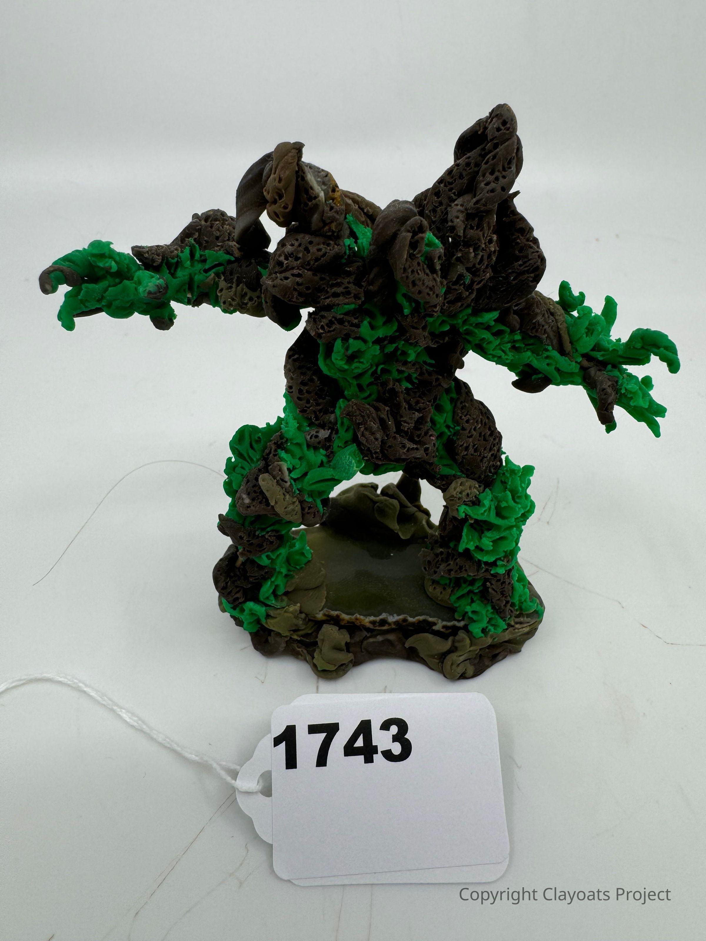 The image portrays a detailed sculpture of a creature made from textured materials in shades of green and brown, resembling a blend of natural elements like wood and leaves. The figure stands in a dynamic pose on a rocky base, displaying intricate details and organic shapes. The background is plain, drawing focus to the sculpture's complex design and earthy color palette. The structure conjures an essence of mythical or fantasy themes