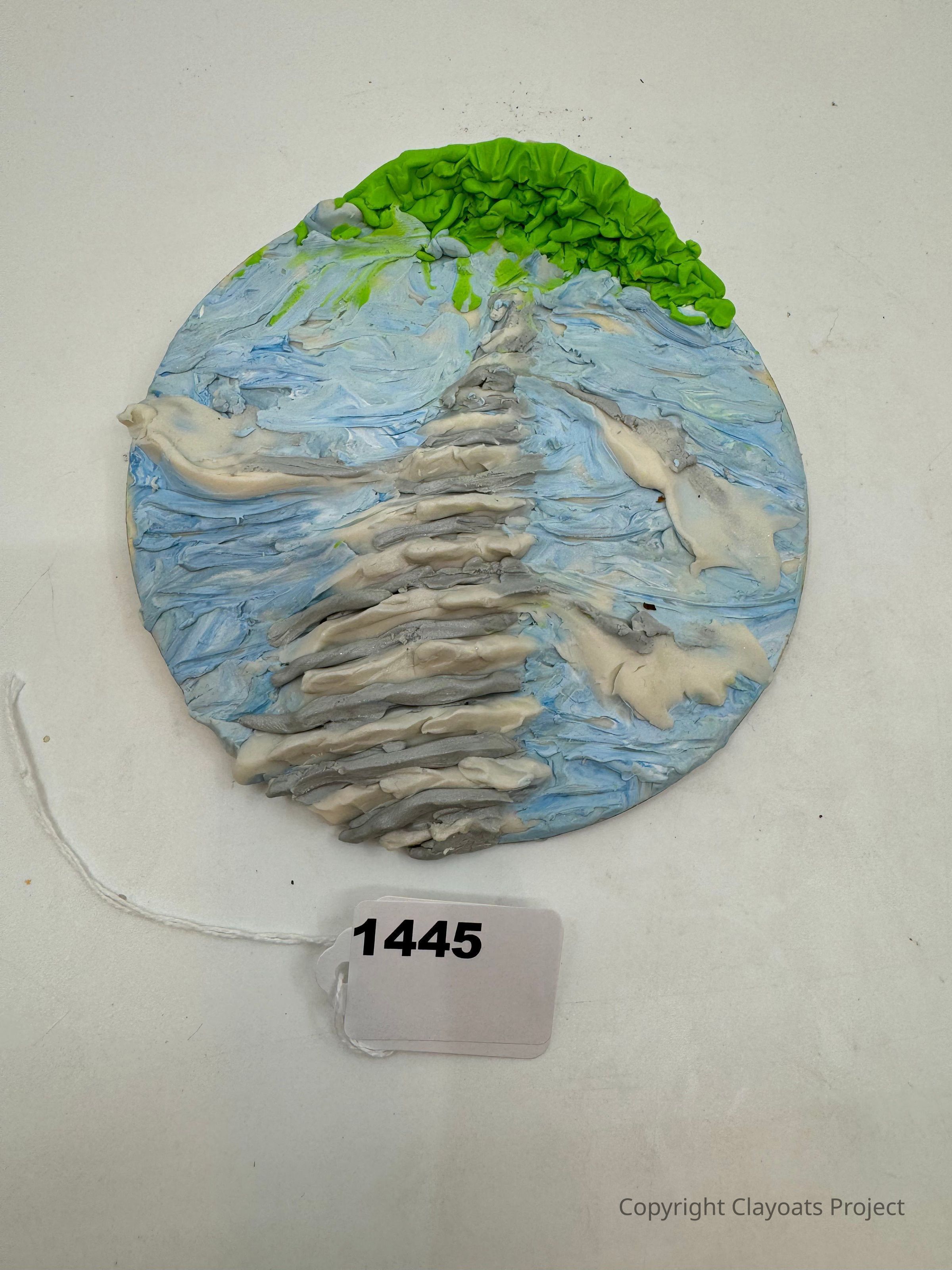 The image features a circular clay artwork with textured layers, resembling a stylized landscape; it includes blue and white colors mimicking a body of water or skies, and vibrant green at the top suggesting vegetation or trees. The rippling effect gives a sense of movement or flow, while a paper tag labeled "1445" is attached, lying on a plain white surface. This combination of colors and textures evokes a natural scene with a three-dimensional quality, encouraging viewers to interpret its artistic representation.