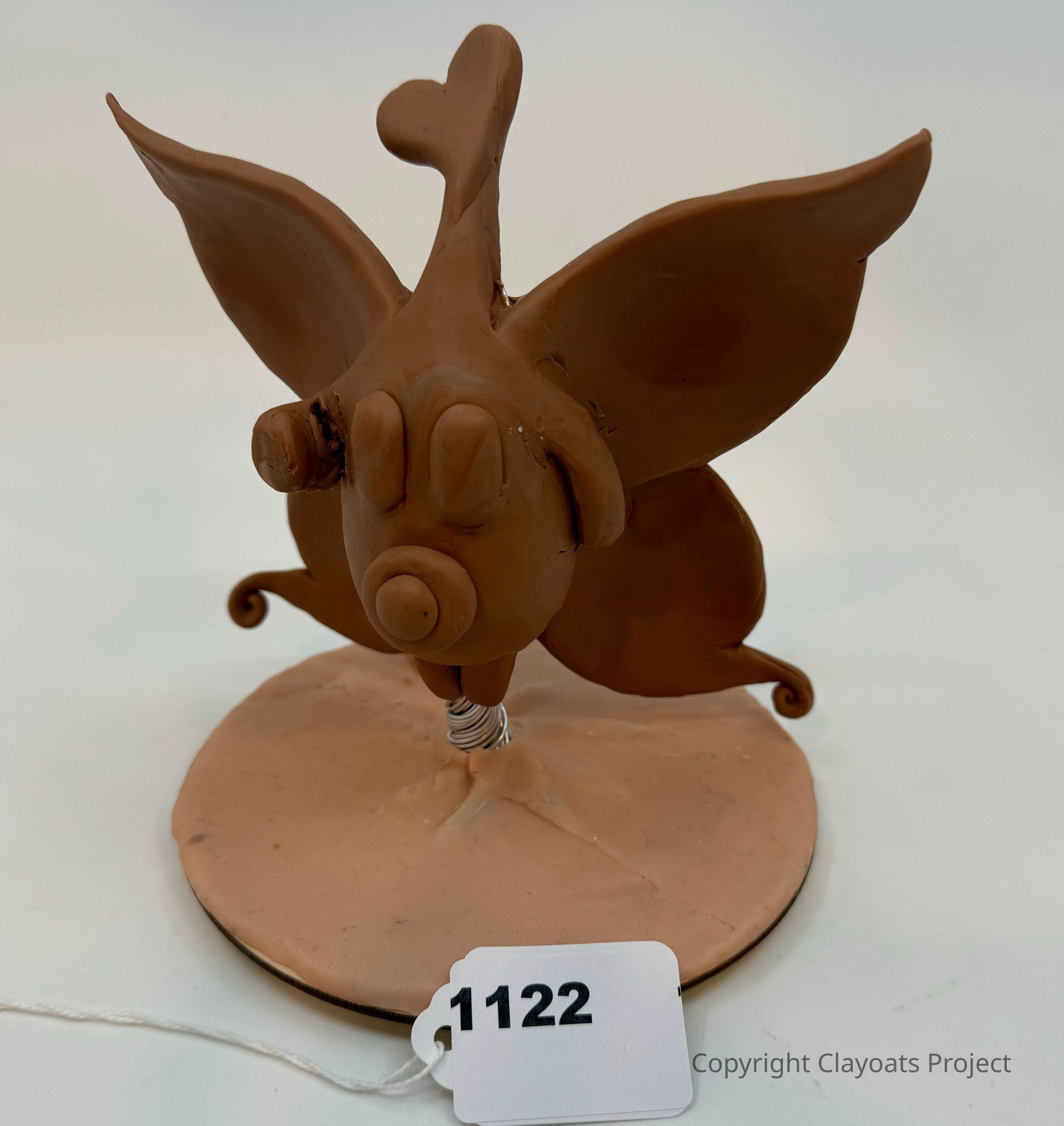 The image depicts a charming clay sculpture of an abstract creature, characterized by large, wing-like ears and a whimsical, elongated snout ending in a circular protrusion. This figure stands on a circular base with a wire support visible beneath, suggesting an imaginative, playful form of art. The sculpture's earthy tone adds to its organic, handcrafted appearance, capturing an essence of