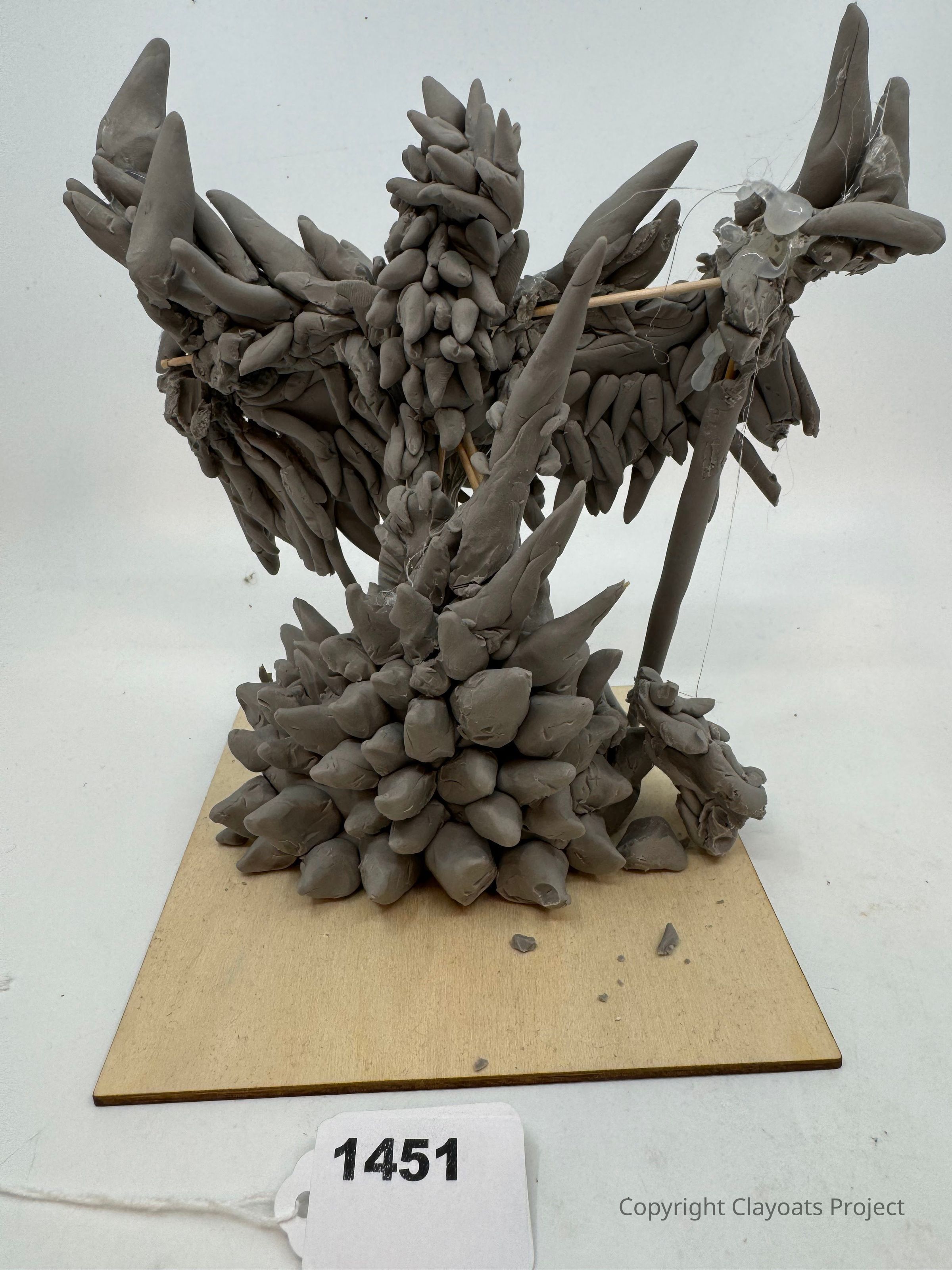 The image features an abstract, spiky sculpture made of gray clay, positioned on a square wooden base showcasing multiple protrusions that create a jagged, dynamic form, resembling organic or crystalline growths in a raw, unfinished state, with visible texture and irregular shapes highlighting the handcrafted nature of the artwork, casting subtle shadows on the surface, and exhibiting a unique aesthetic that draws viewers' attention to its intricate, layered composition.