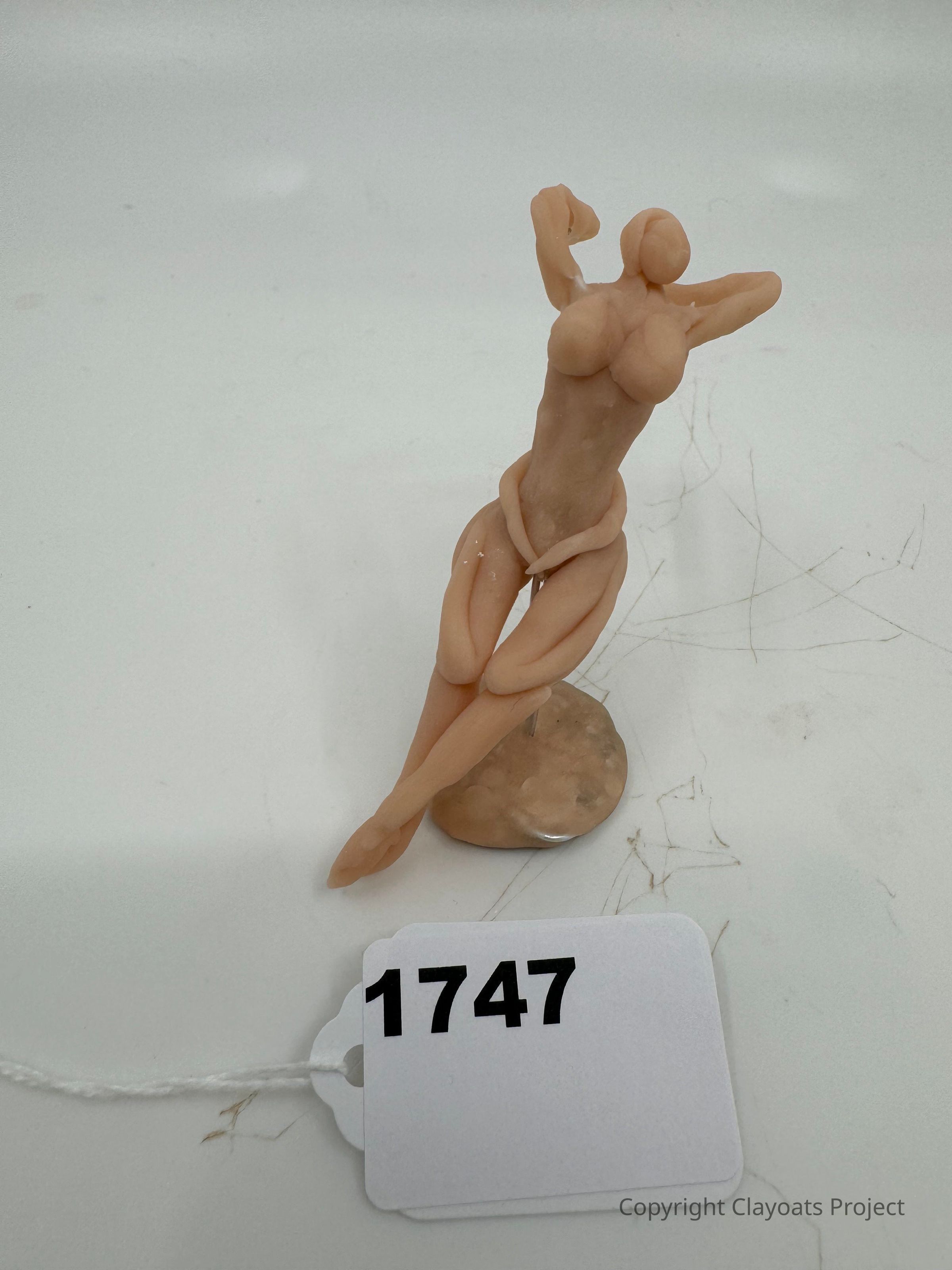 The image features a small, abstract clay sculpture of a human figure in a relaxed pose, with its arms raised above its head and legs crossed, standing on a circular base. The figure is sculpted in a simplistic style, emphasizing the human form without detailed features. The sculpture is placed on a flat surface,. The background is plain, highlighting the artwork. The soft, neutral color of the clay contrasts subtly with
