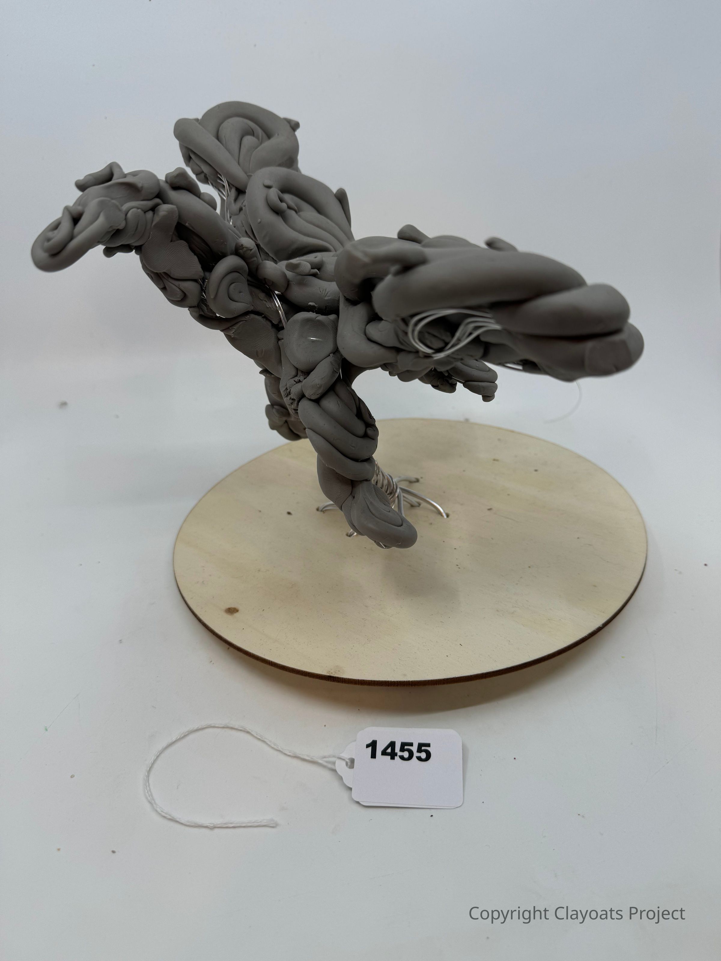 The image displays a sculptural piece made of grey clay, intricately twisted and shaped into a dynamic, abstract form that seems to suggest movement or flight, mounted on a circular wooden base; while the sculpture itself is presented against a plain background, emphasizing its unique texture and creative craftsmanship, inviting viewers to interpret its artistic intent and form through its complex, imaginative design