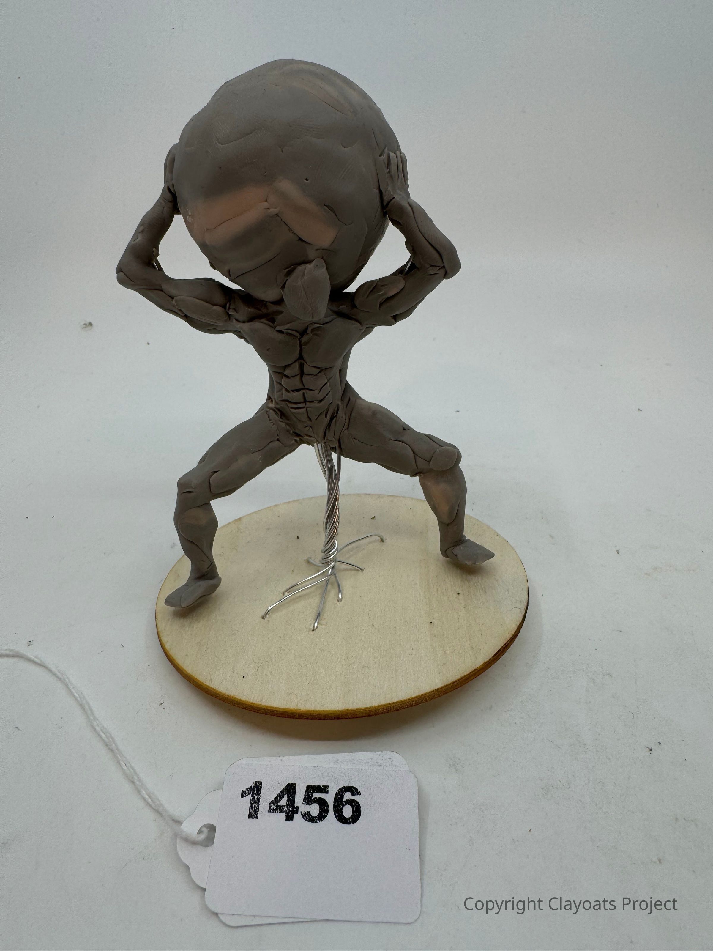 The image depicts a sculpture of a muscular figure with defined anatomy, crouched with both hands lifted above its head, supporting a large, spherical object. The sculpture is mounted on a circular wooden base with wire support visible beneath. The number "1456" is printed on a white paper label, attached to the base by a piece of string. The figure's pose is reminiscent of the mythological character Atlas, often depicted bearing the weight of the world. The white background provides a neutral setting, emphasizing