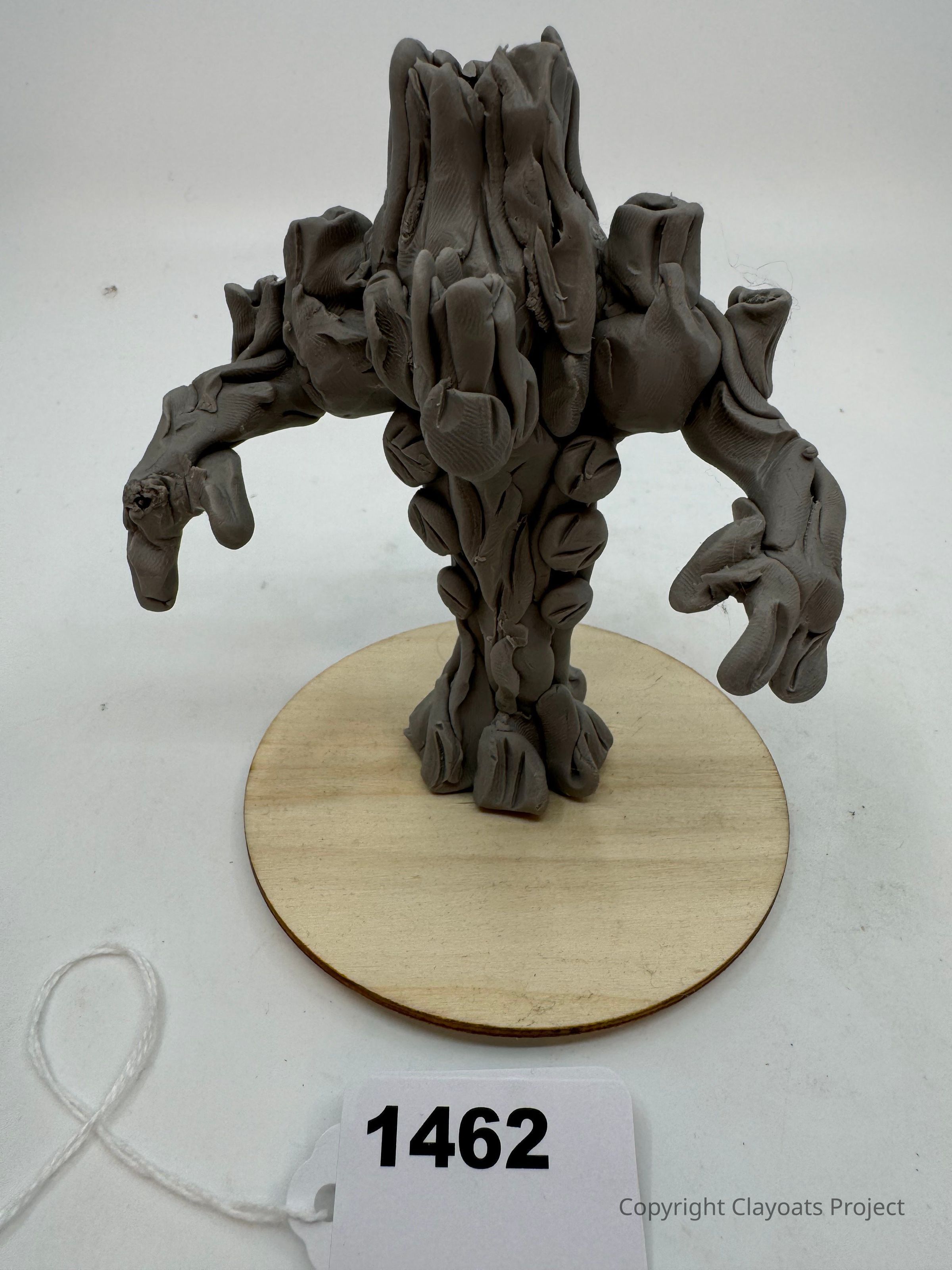 The image depicts a creatively sculpted clay figure resembling a tree-like creature with a robust and textured surface, standing on a circular wooden base. Its intricate details, including twisted branches and rugged bark texture, add depth to its appearance. The figure exhibits outstretched arms and a solid trunk, giving an impression of strength and rootedness. The sculpture's earthy tone enhances its natural, organic look.