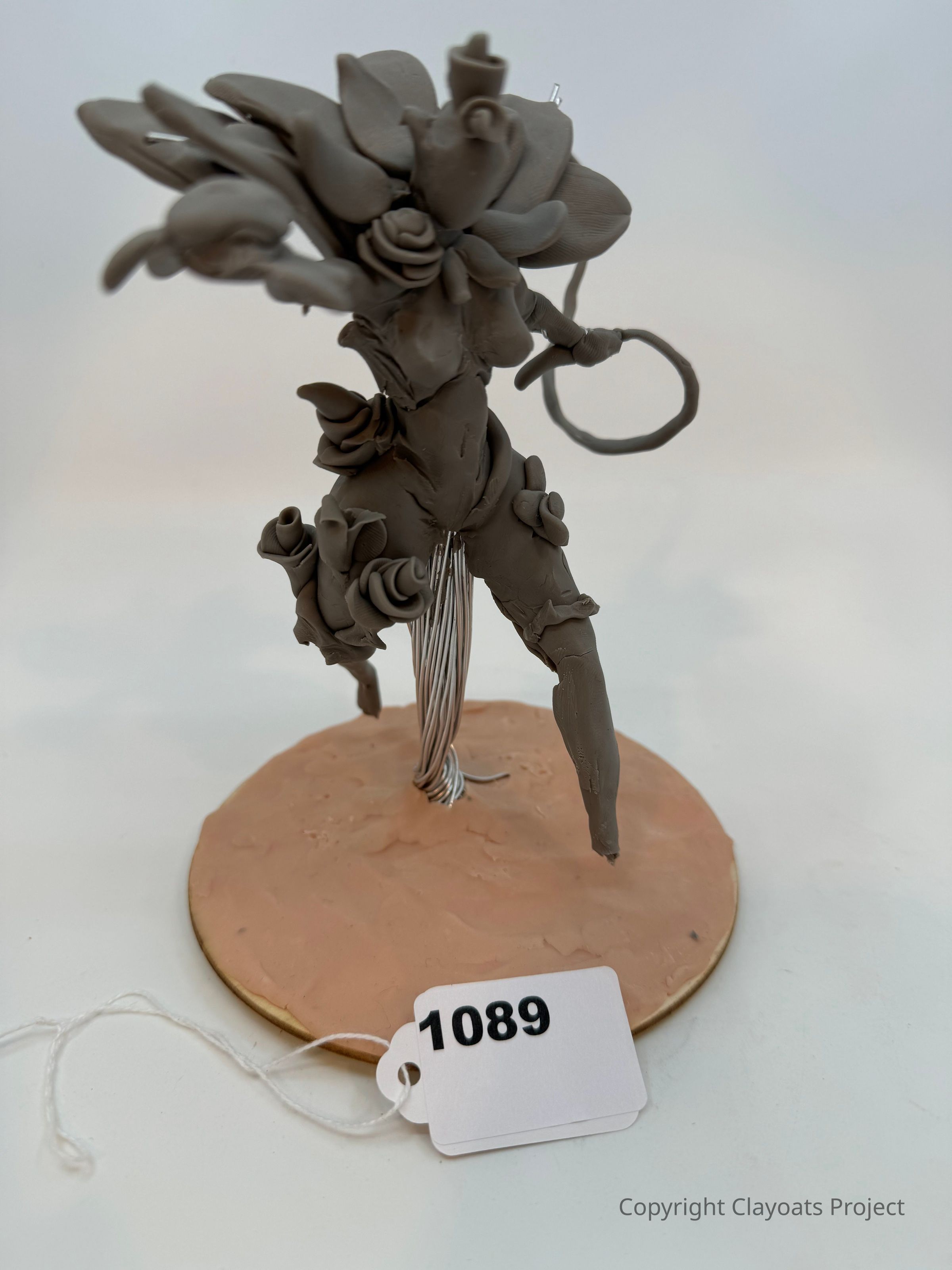 The image shows a detailed clay sculpture of a humanoid figure that appears to be intertwined with organic elements such as leaves or petals, mounted on a circular base. The figure's limbs and body are adorned with intricate swirls and curls, suggesting a fusion of natural and fantastical elements. The sculpture's textured surface and dynamic pose convey a sense of movement and fluidity, while the neutral clay color highlights the intricate