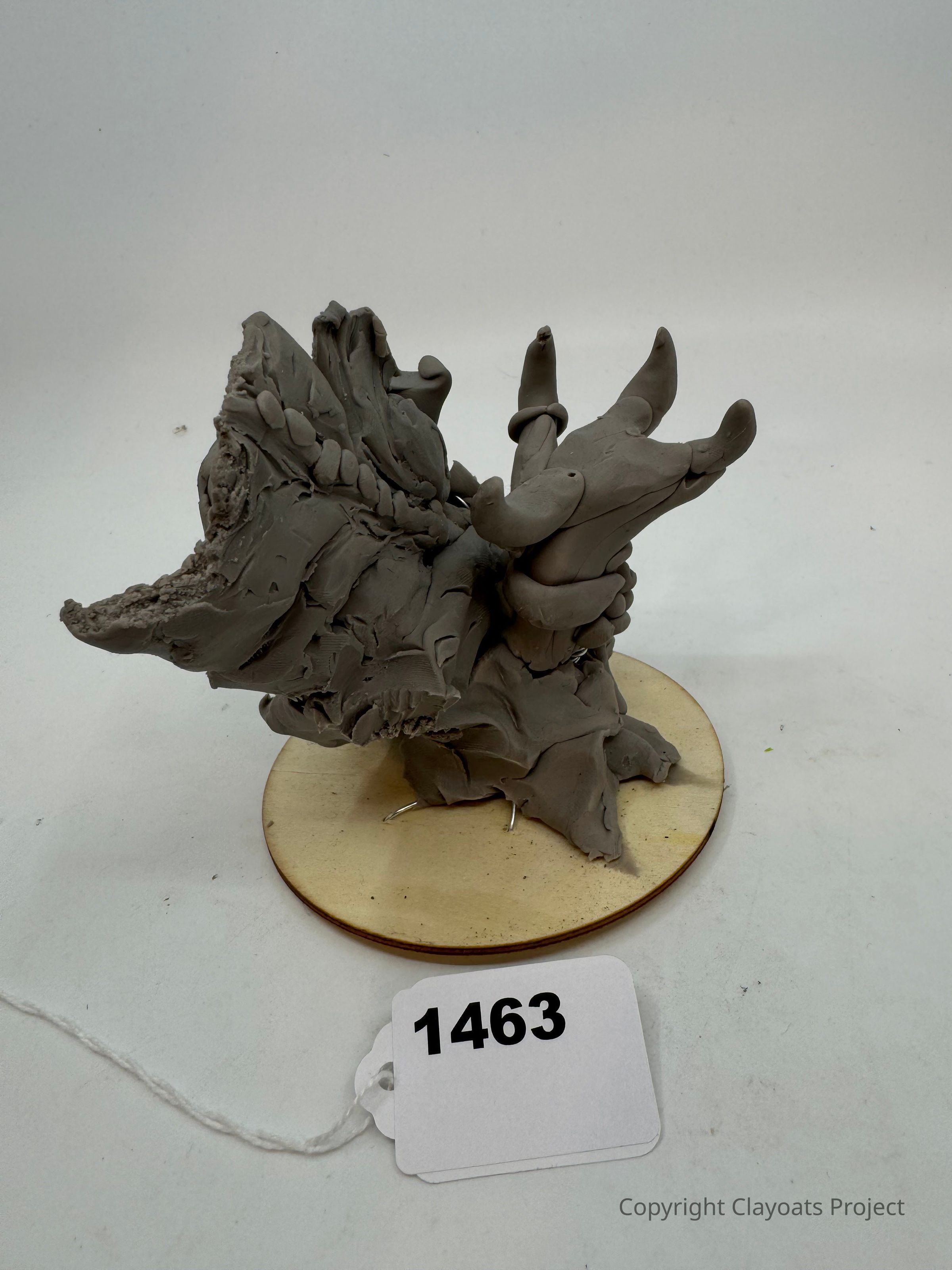 The image features a gray, abstract sculpture mounted on a circular wooden base, depicting an intricate, organic form with multiple curved and pointed elements. The piece has a rough texture, suggesting a natural earthy feel. The arrangement exudes a sense of motion and dynamism, as if the structure is captured mid-transformation, showcasing artistic creativity in its design.