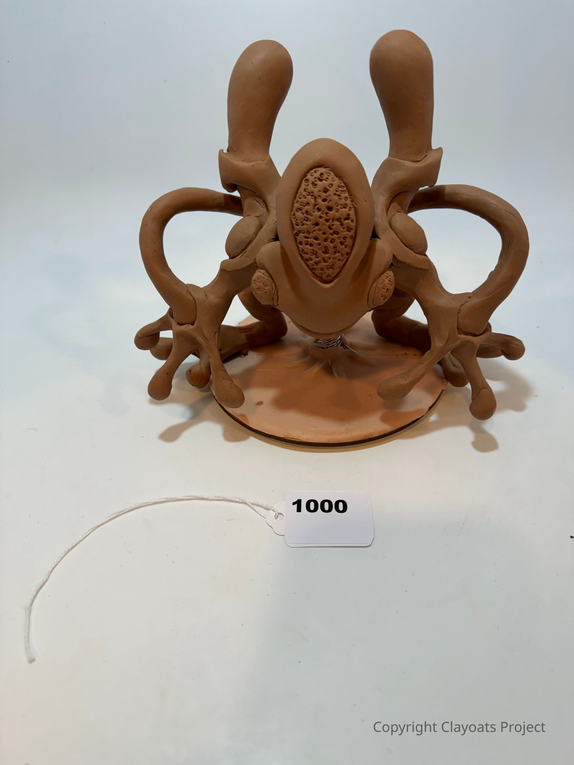 The image features a sculptural piece resembling an abstract or alien creature, crafted from a brown, clay-like material with intricate details, such as textured surfaces and multiple limb-like extensions; its form suggests a mixture of organic and geometric elements, creating an intriguing, otherworldly appearance that stimulates curiosity and imagination. The sculpture sits on a flat base, accompanied by a numbered tag reading "1000," indicative of its cataloging or collection status, suggesting that it might be part of an exhibition or inventory.